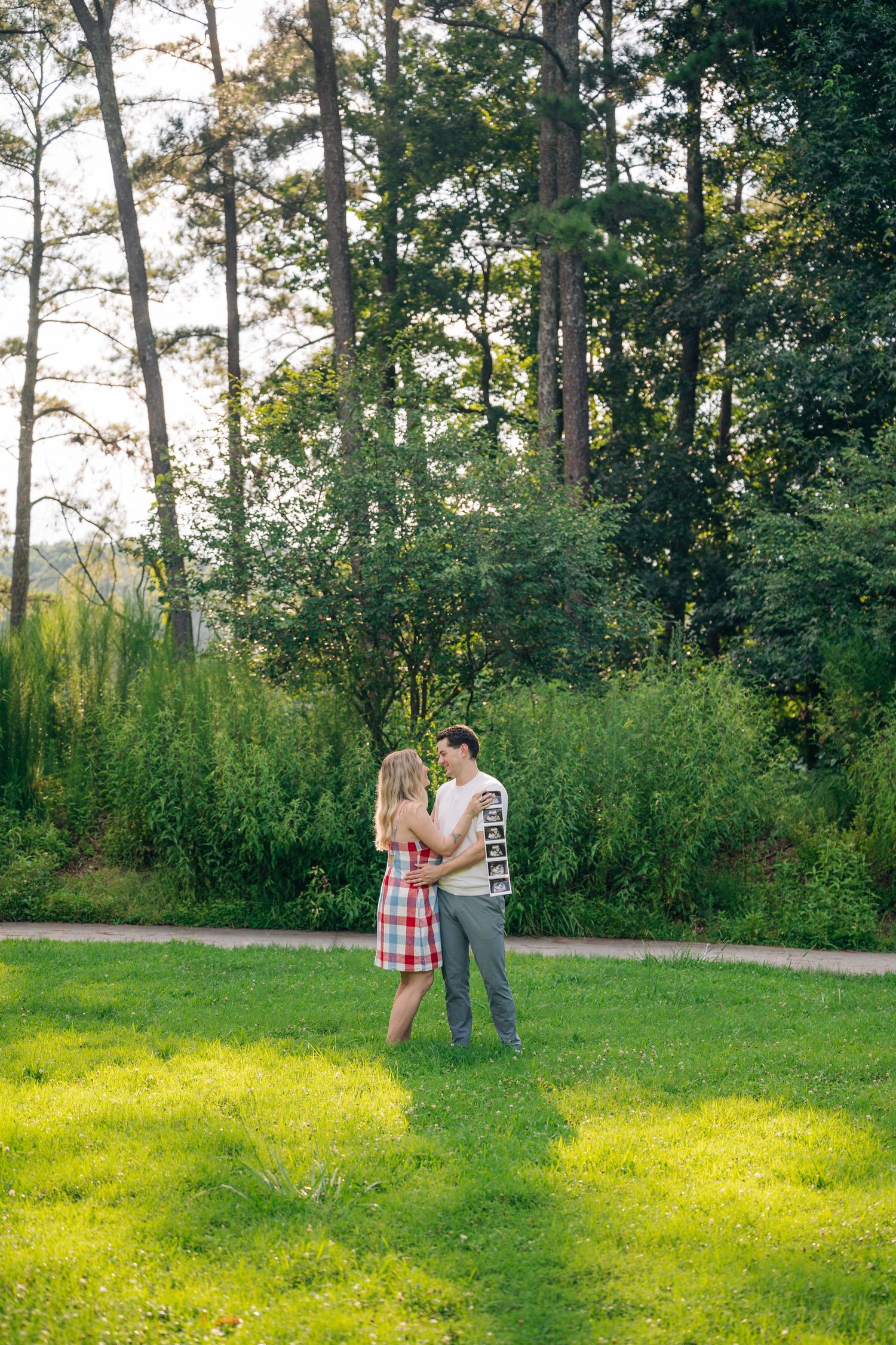A couple embraces on a green lawn surrounded by lush trees and shrubs in a sunny park setting.