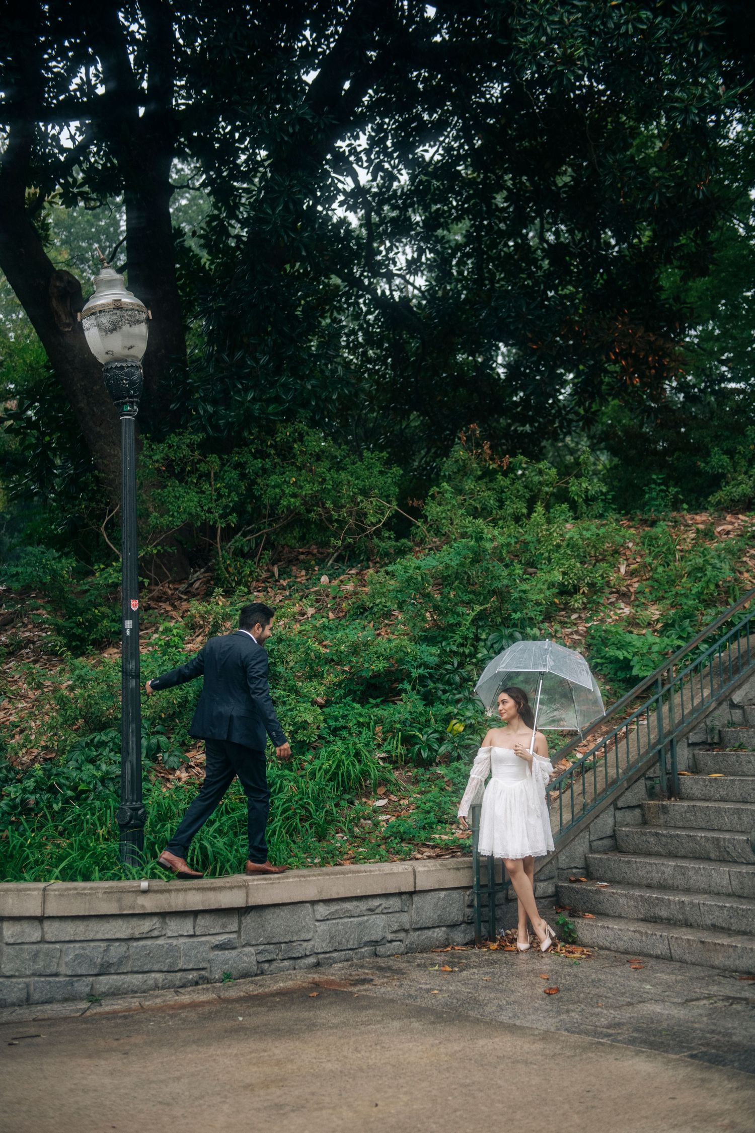 Bride in white dress holds clear umbrella on stone steps surrounded by lush green park.