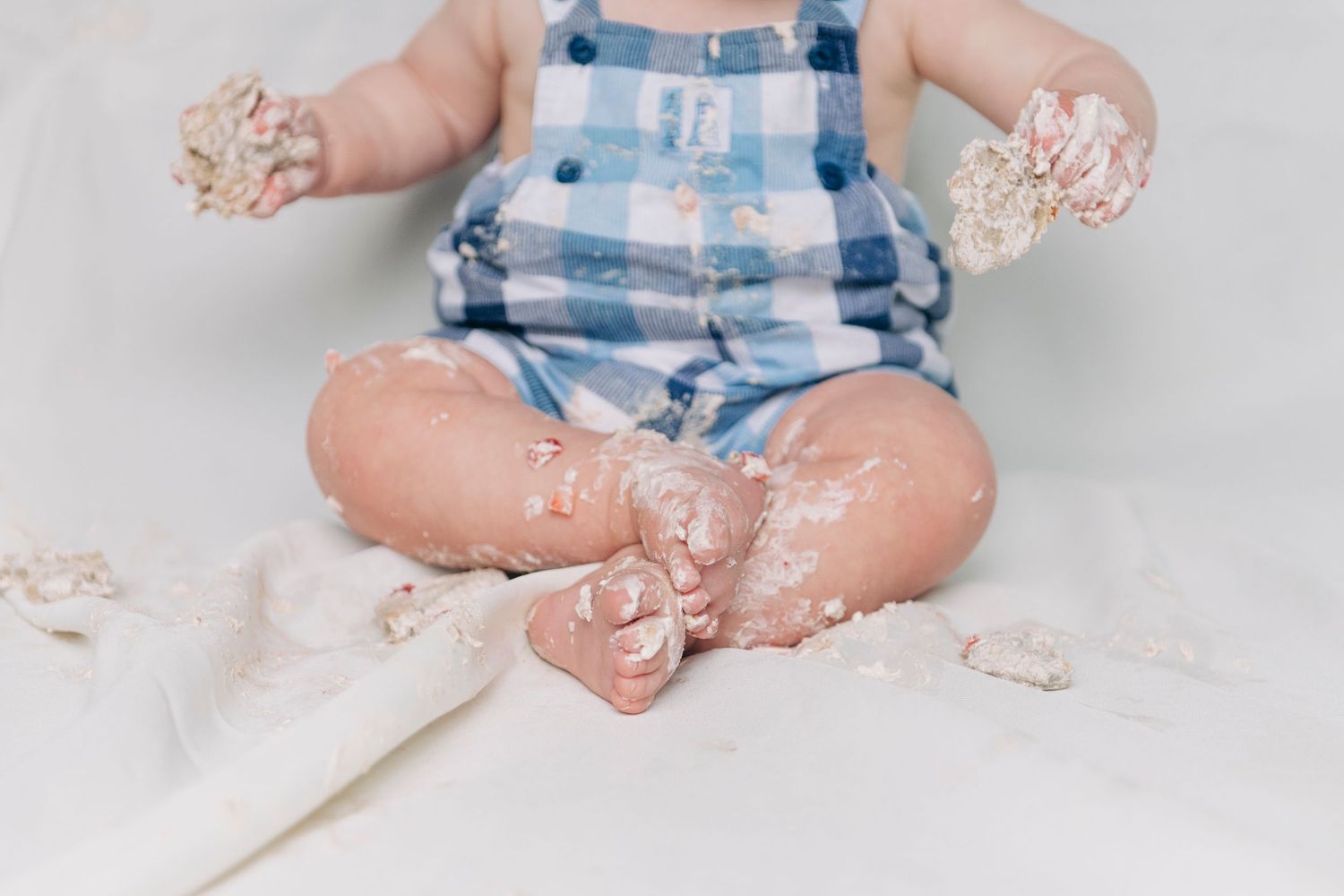 Baby in blue plaid overalls sits on white backdrop with cake smash mess on hands and feet.