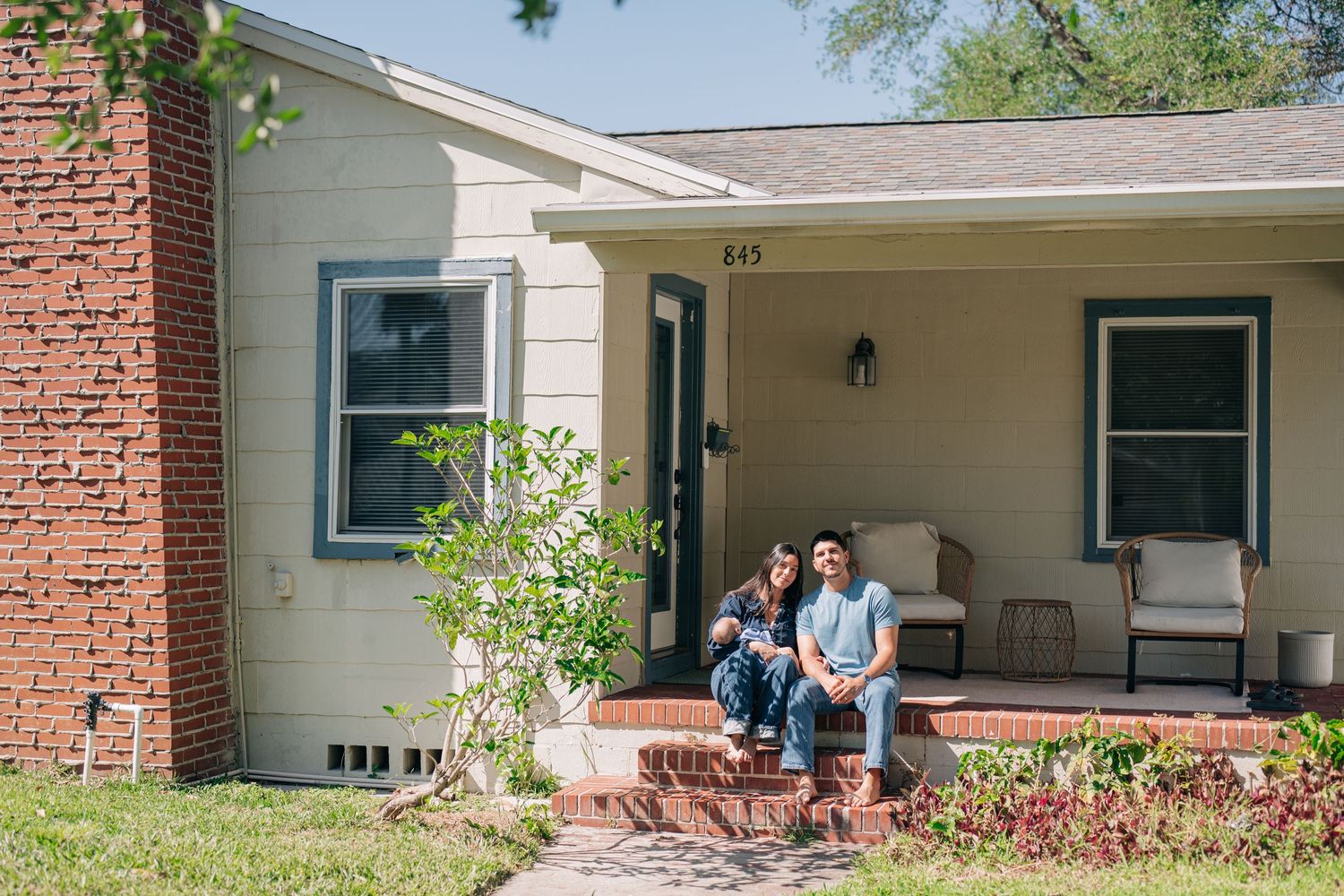 Couple sitting on brick steps of charming ranch-style home with teal trim and covered porch.