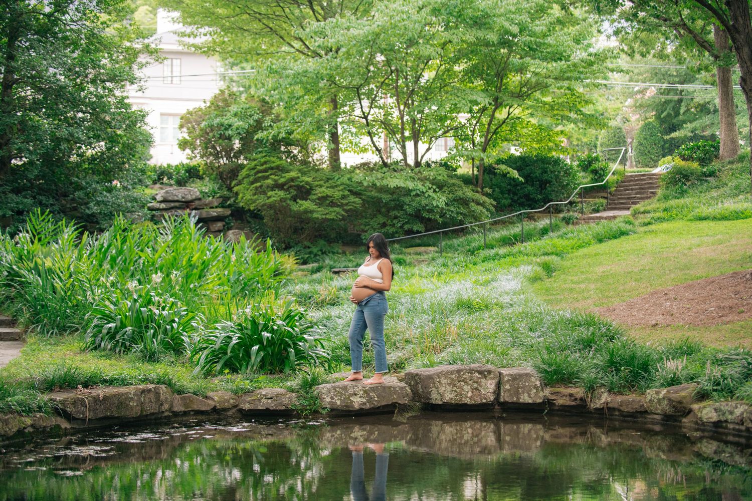 A woman stands on rocks beside a serene pond in a lush green garden park surrounded by trees and wildflowers.