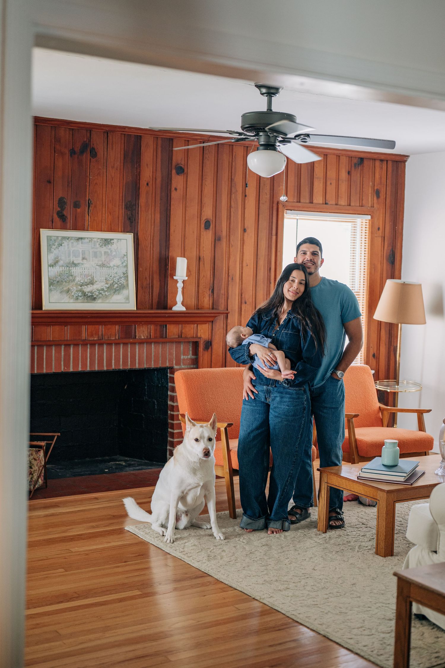 Young couple holding newborn baby with white dog in cozy wood-paneled living room with brick fireplace.