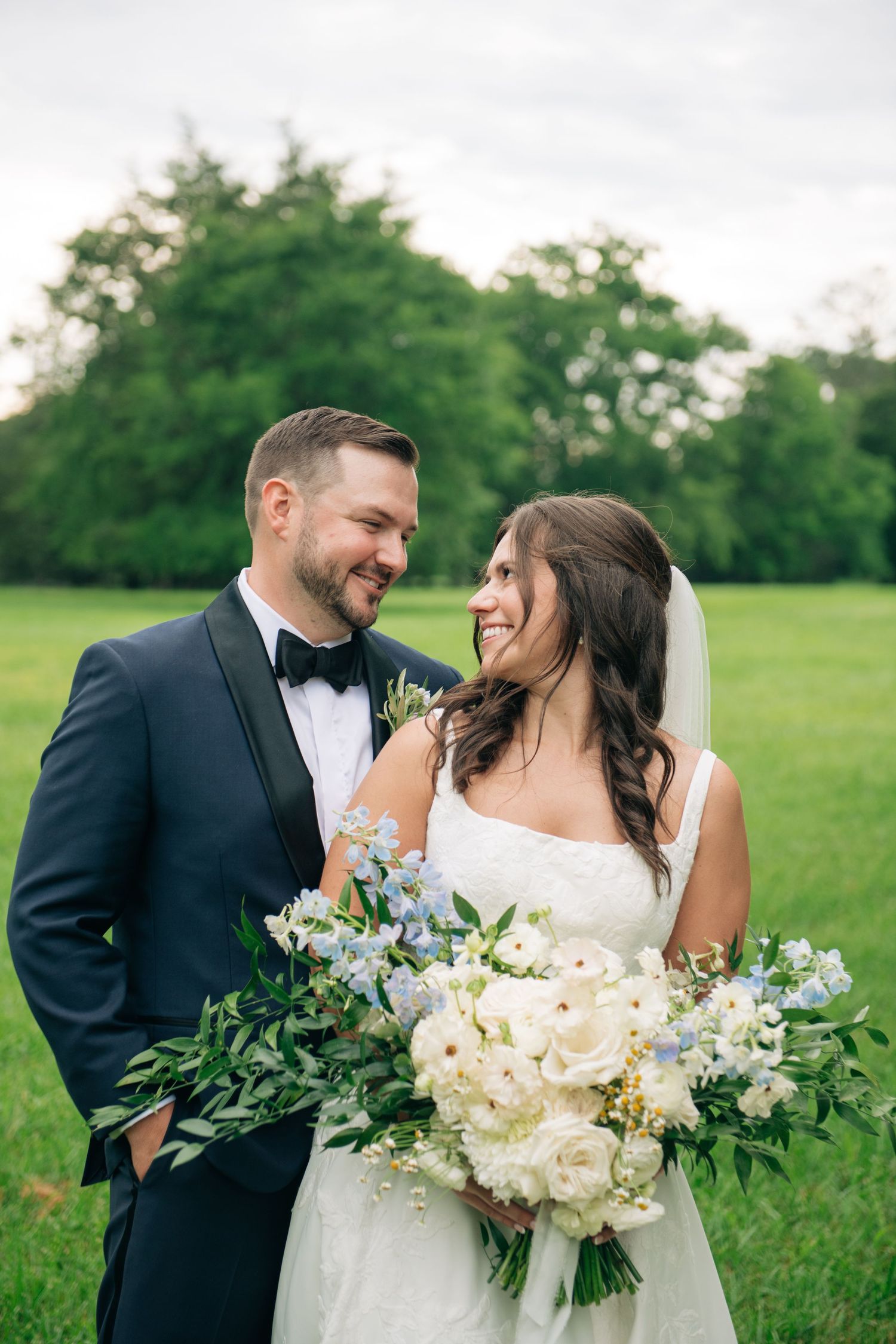 Smiling bride and groom in navy tuxedo share a romantic moment outdoors, bride holding white and blue floral bouquet.