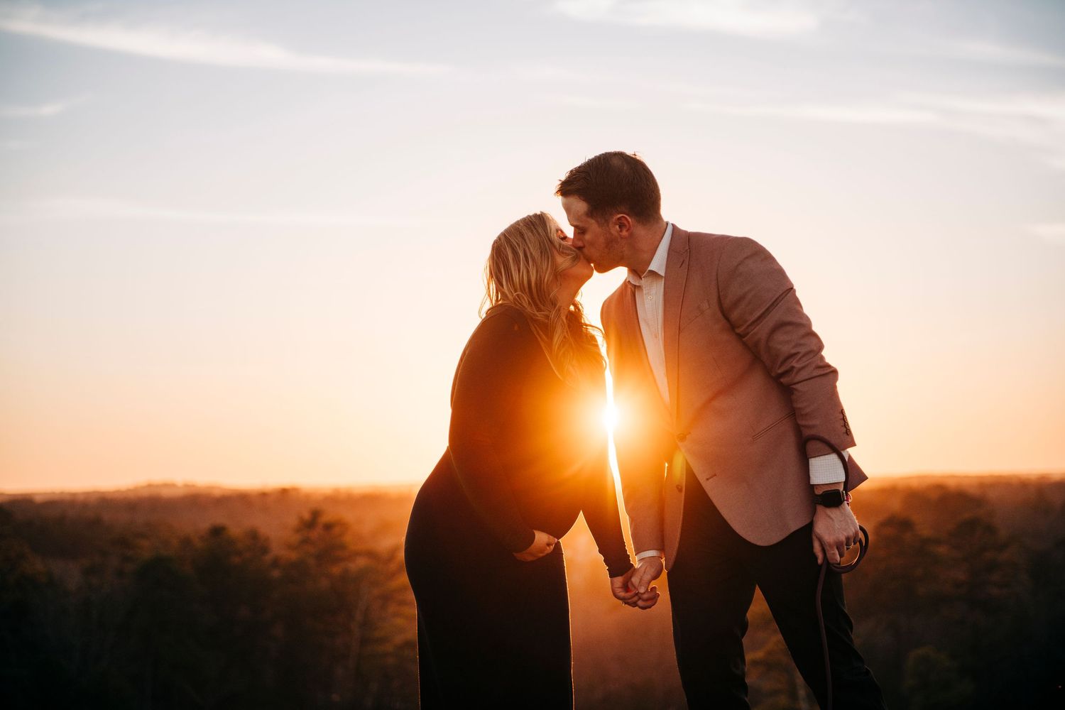 Couple sharing a romantic kiss at golden hour sunset, holding hands outdoors with warm glowing light behind them.