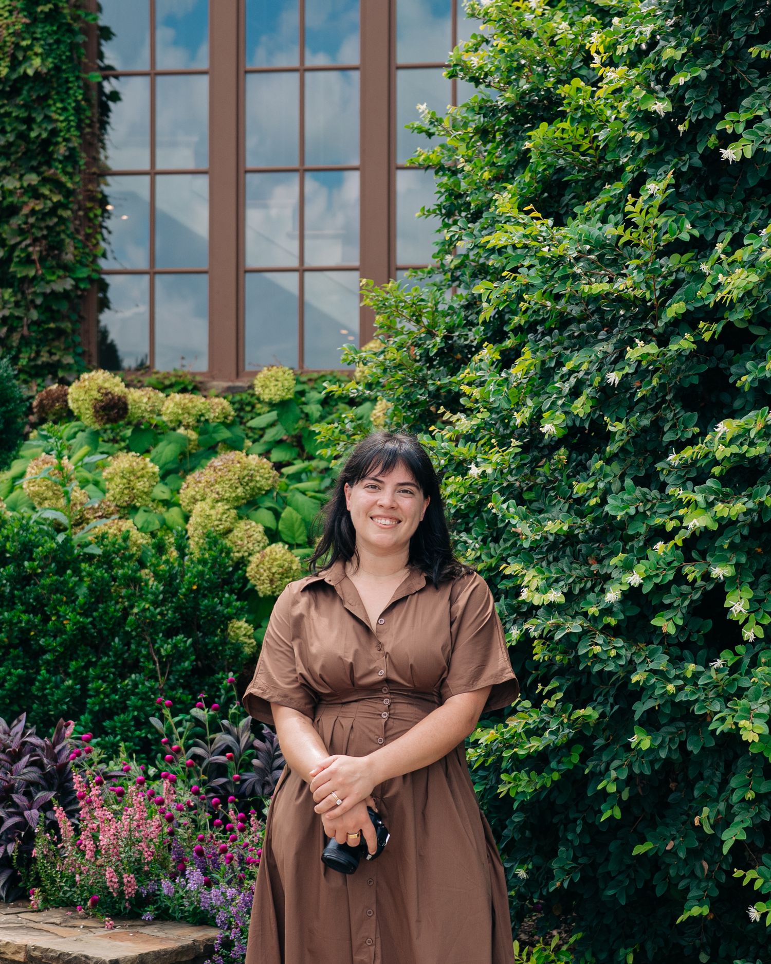 Woman in brown dress smiling in lush garden with hydrangeas and ivy-covered building.