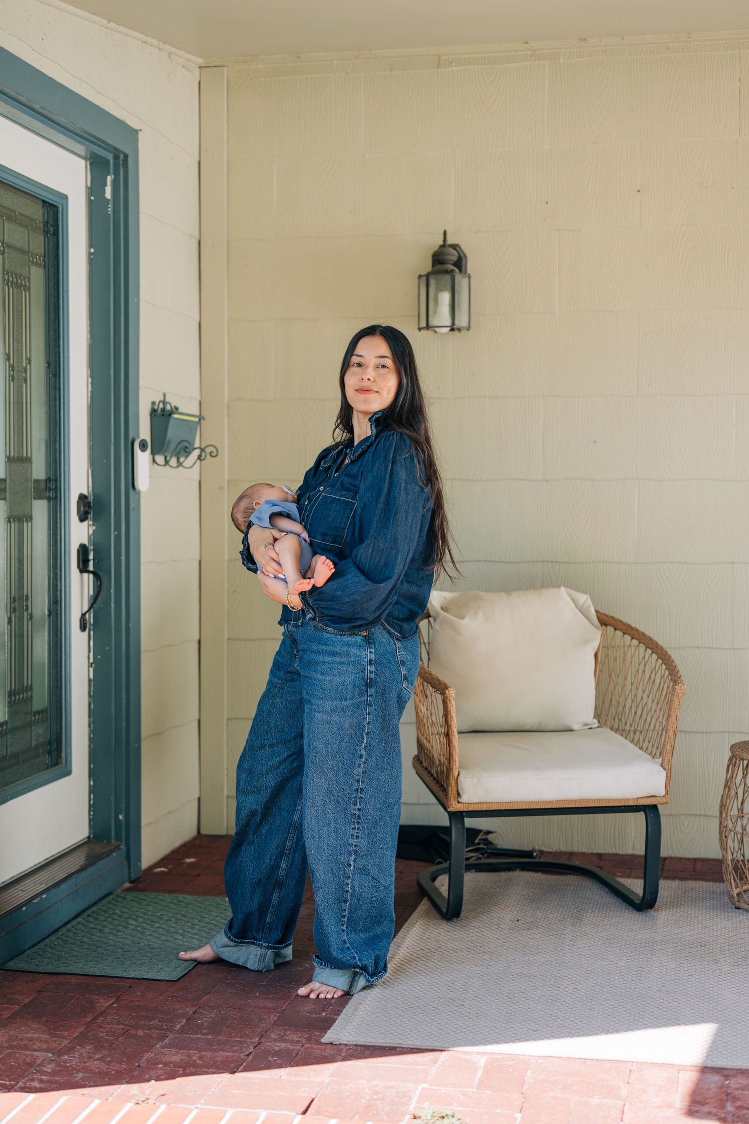 Woman in denim outfit holding newborn baby on a sunlit porch beside a wicker chair.