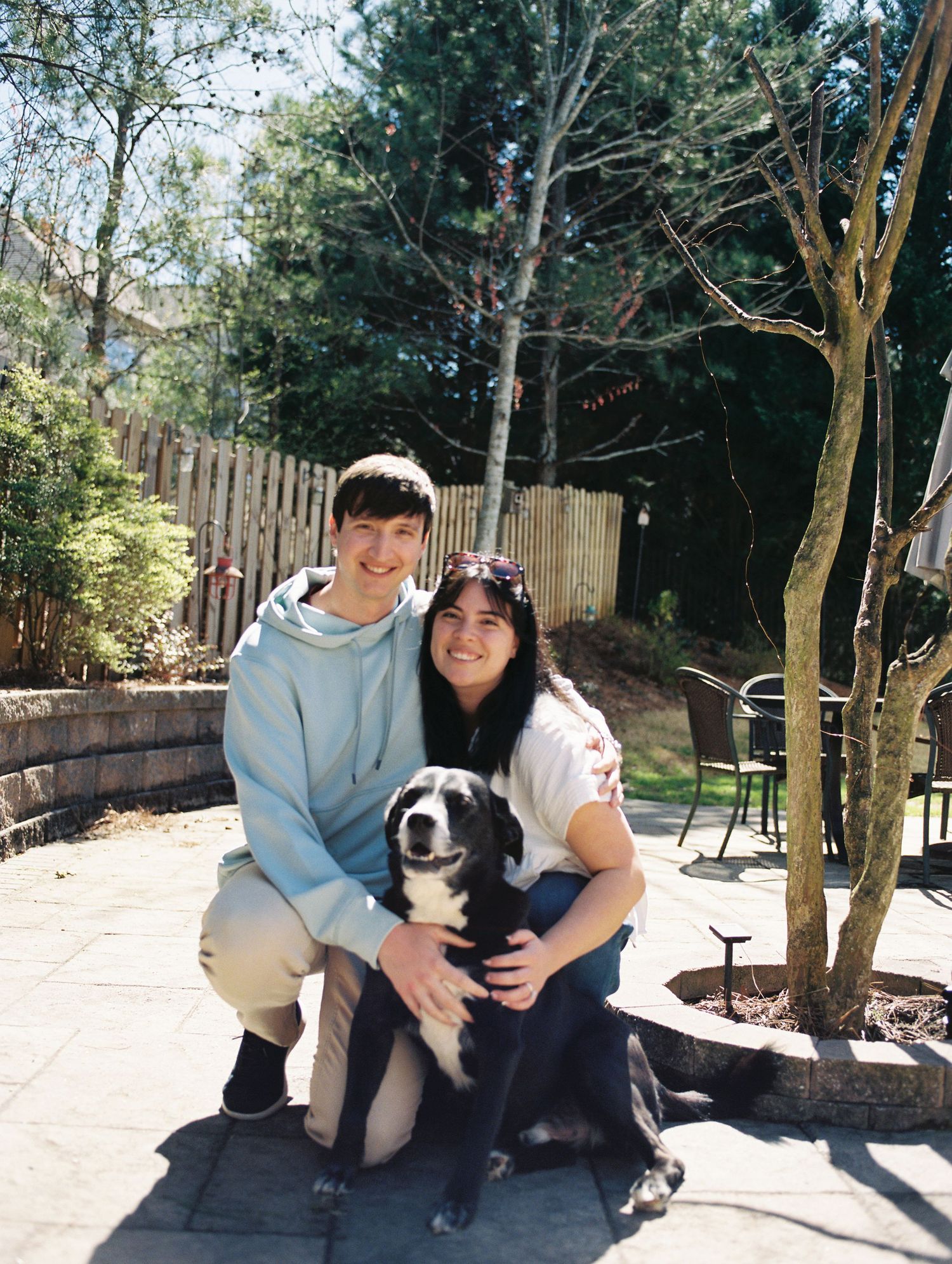 Smiling couple crouching with black and white dog on sunny patio with wooden fence.