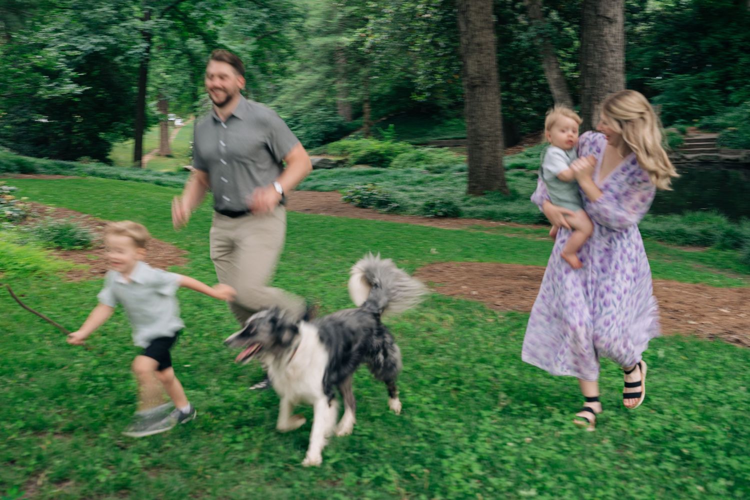 Family with two young children and a fluffy dog playing outdoors in a lush green park on a sunny day.