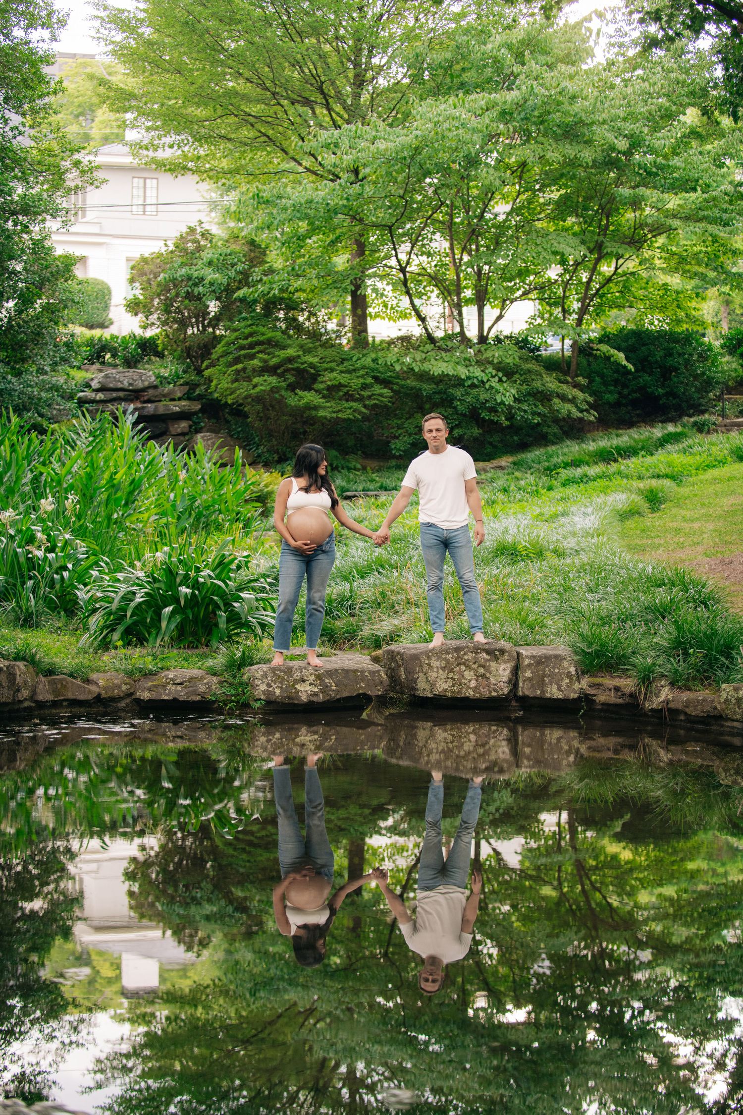 Pregnant couple holding hands on stone bridge over reflective pond in lush green park setting.
