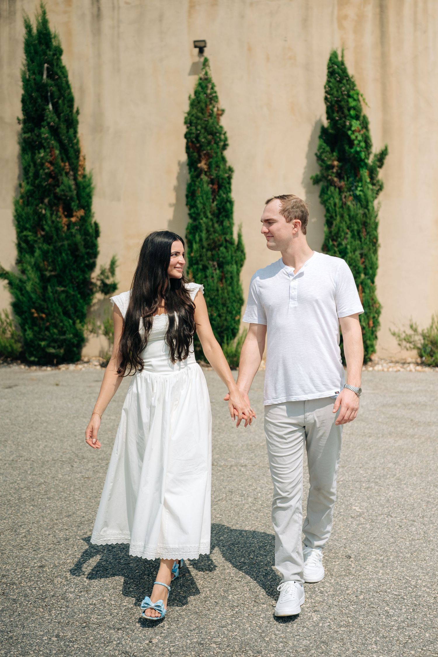 A couple holding hands and smiling at each other while walking outdoors near tall cypress trees and a beige wall.