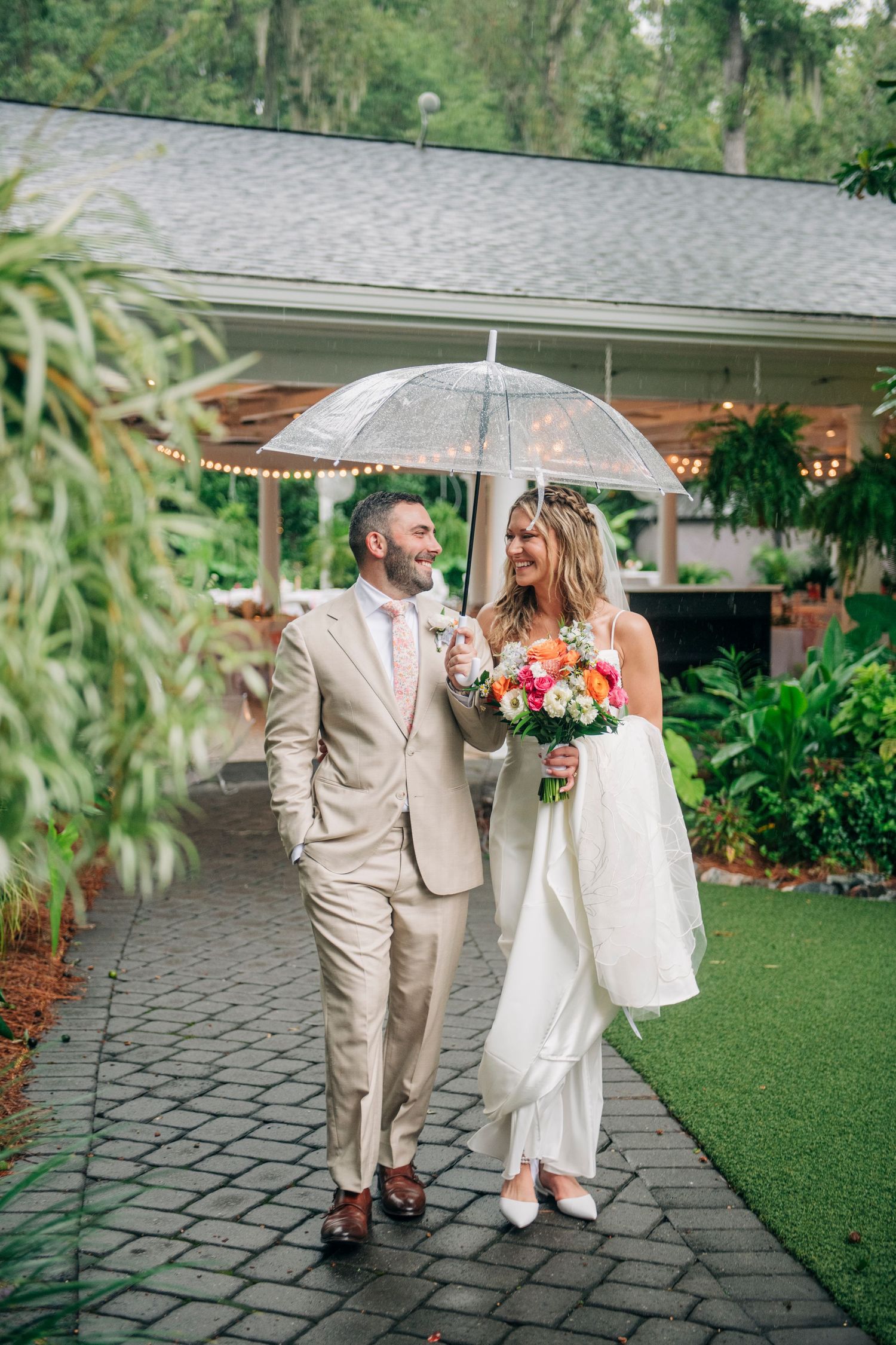 Bride and groom walking under clear umbrella in rain at lush garden wedding venue in Savannah, Georgia, The Mackey house.