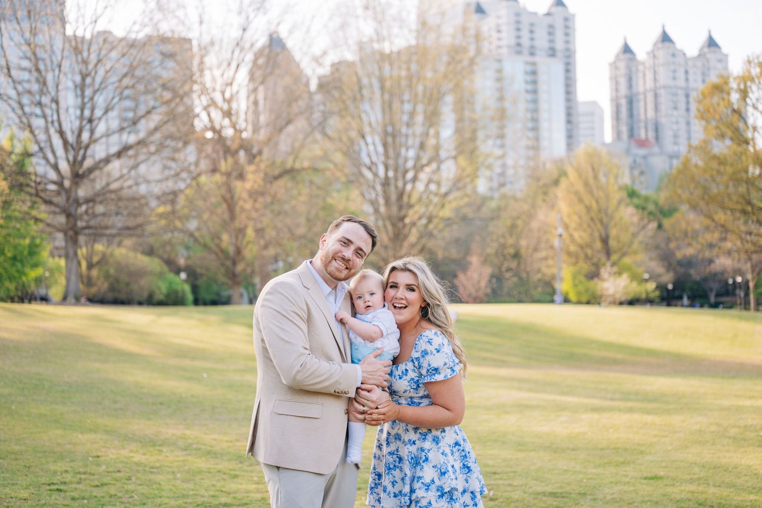 A happy family of three poses in a sunny park with Atlanta city skyline in the background.