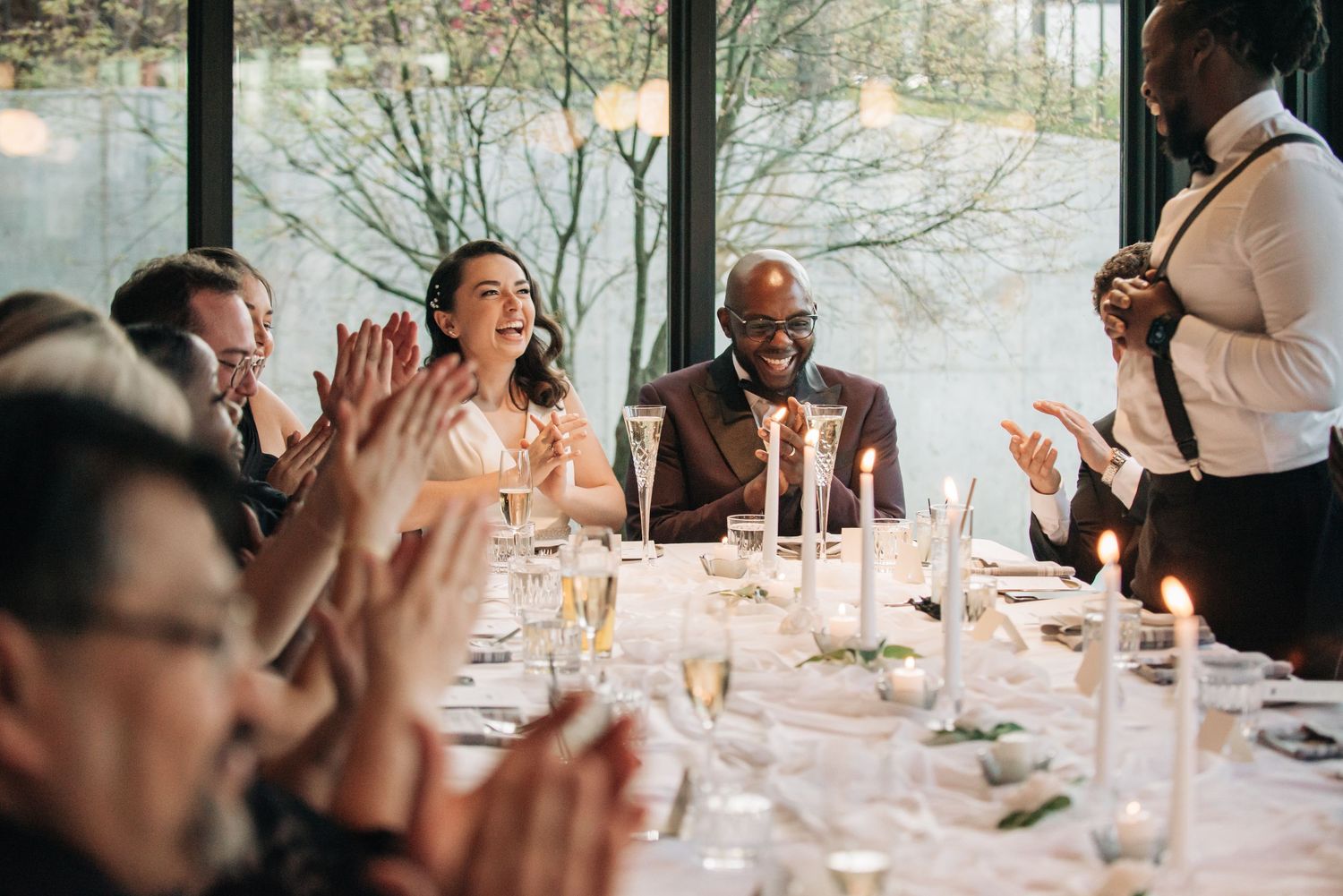 Guests laughing and clapping at an elegant wedding reception dinner table with candles and champagne glasses.