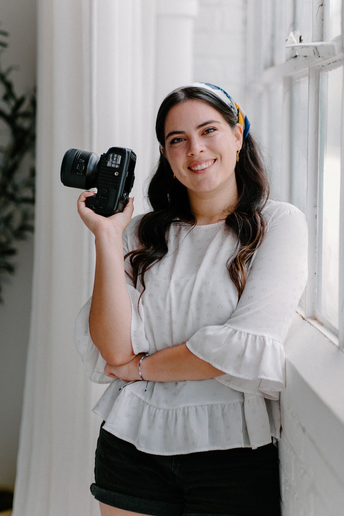 Smiling young woman holding a DSLR camera, wearing a white blouse and colorful headband, posing by a bright window.