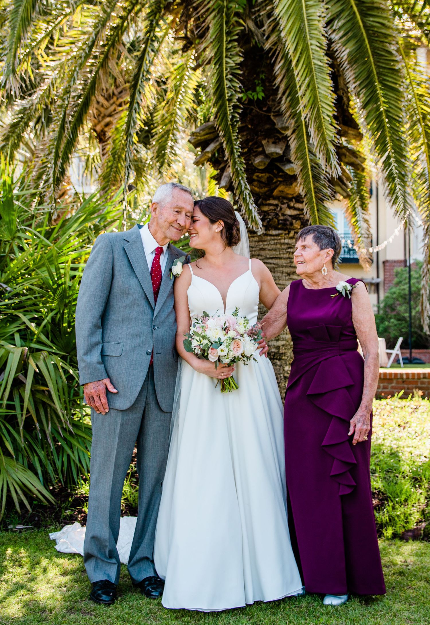 Bride in white gown poses with parents near tropical palm tree, holding pink bouquet at outdoor wedding.