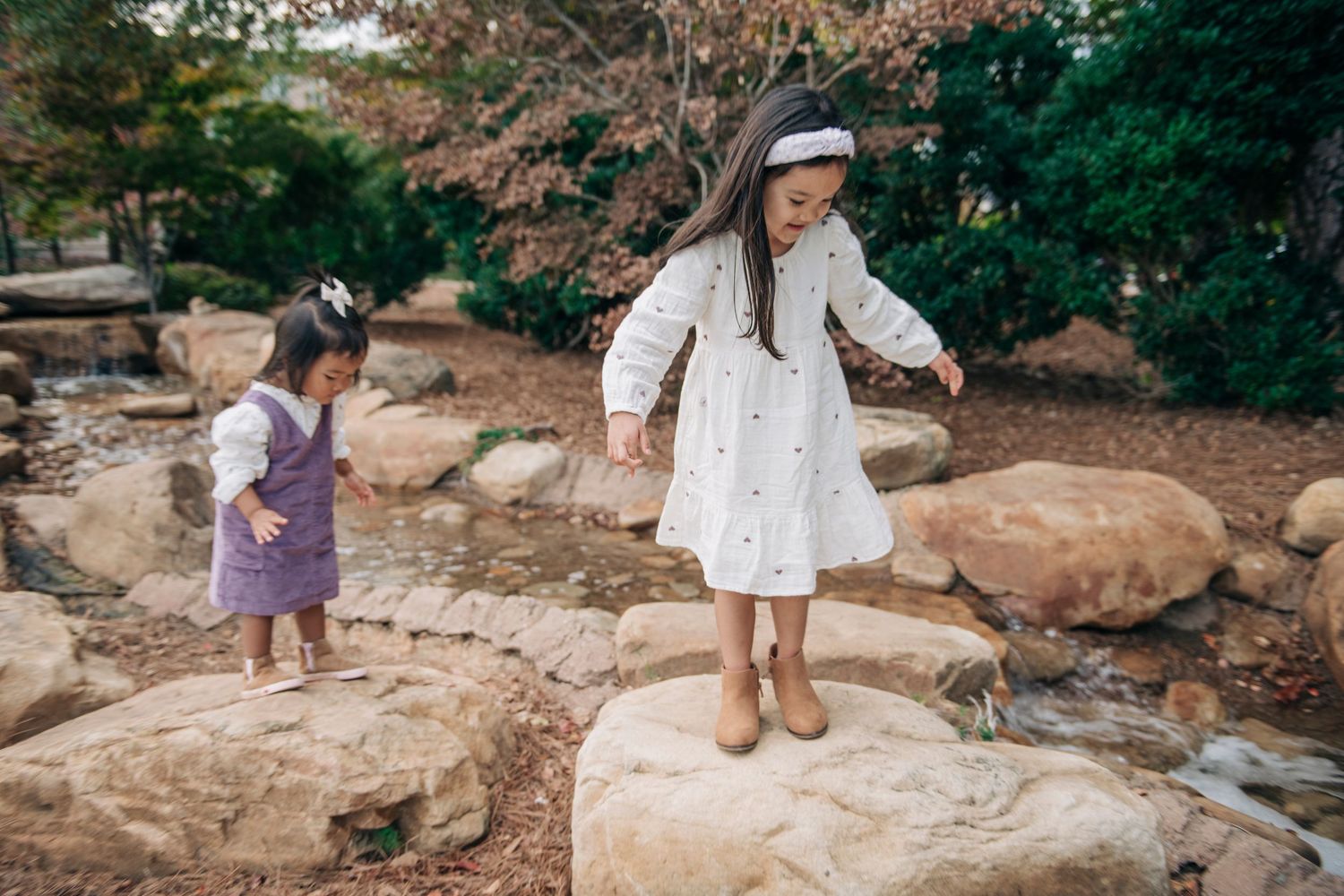 Two young girls in dresses balance on rocks in a scenic garden with lush greenery and a small stream.