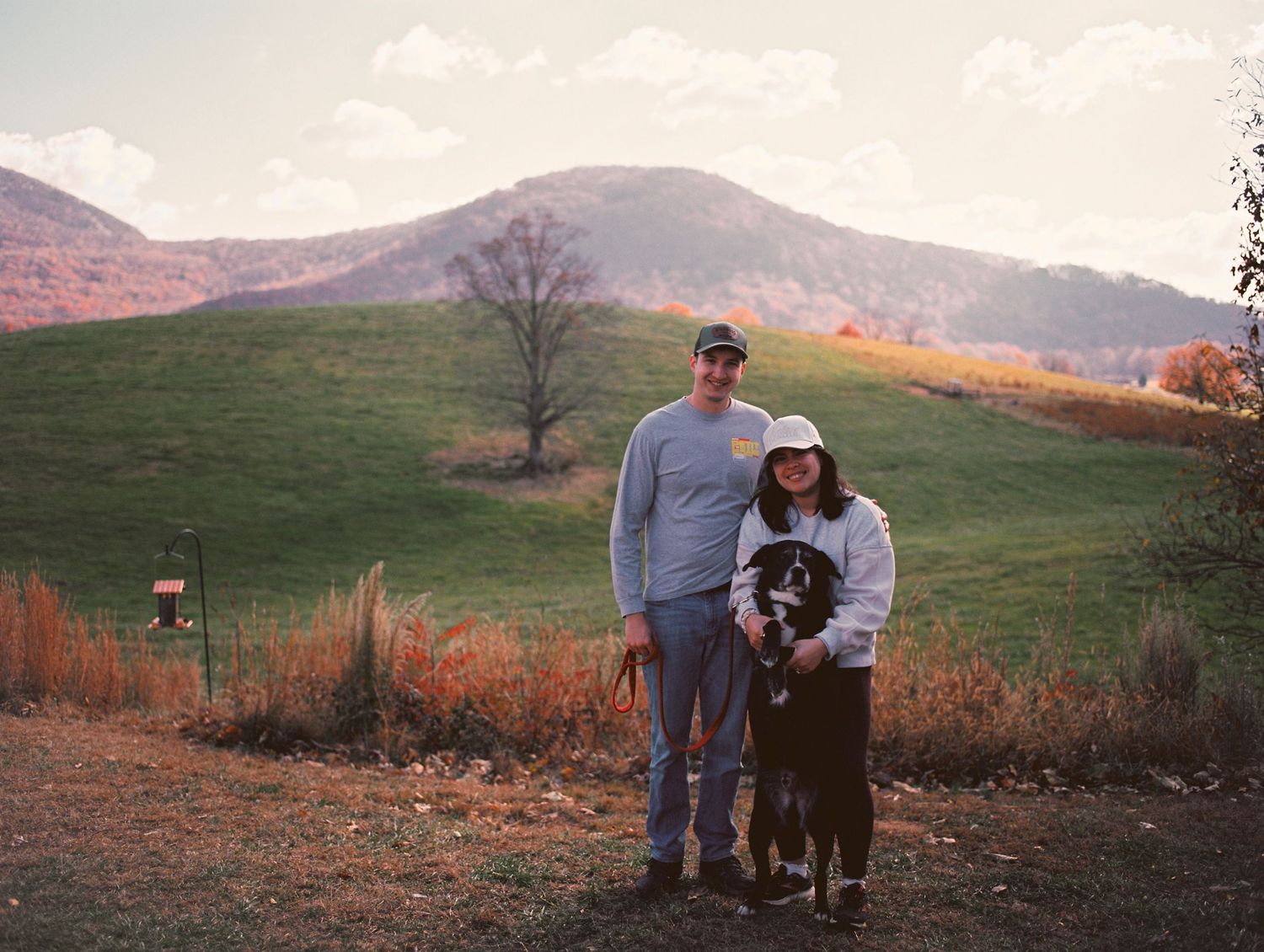 Couple posing with black dog on scenic autumn hillside with mountain backdrop.