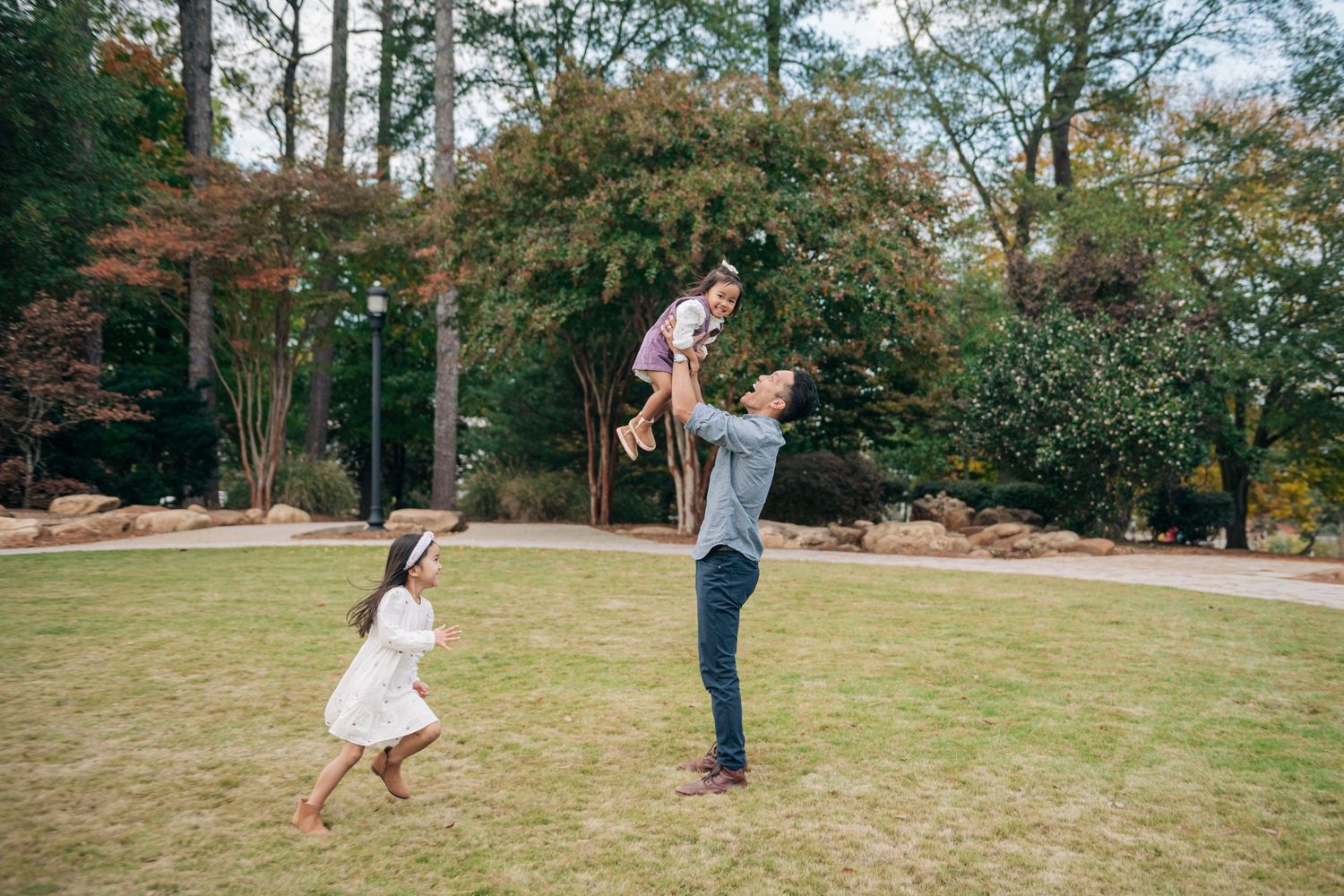 Father joyfully tosses child in air while daughter runs toward them in a lush green park with autumn trees.
