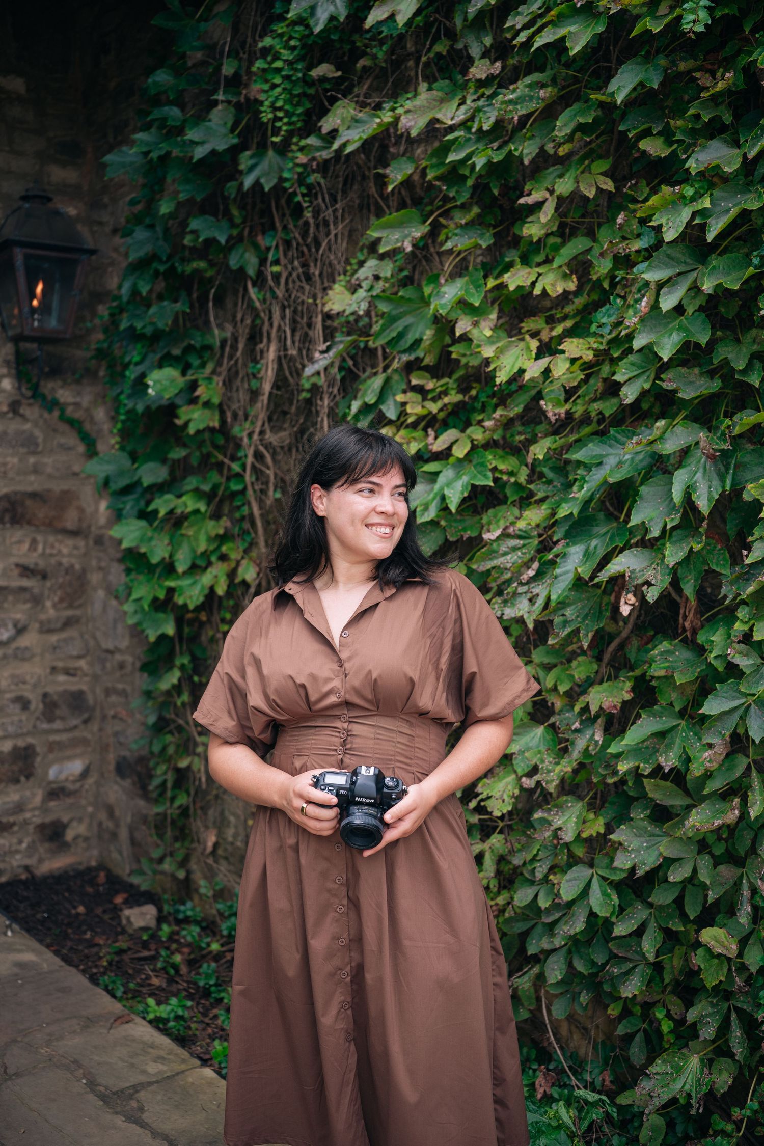 Woman in brown dress holding camera smiling outdoors against ivy-covered stone wall.