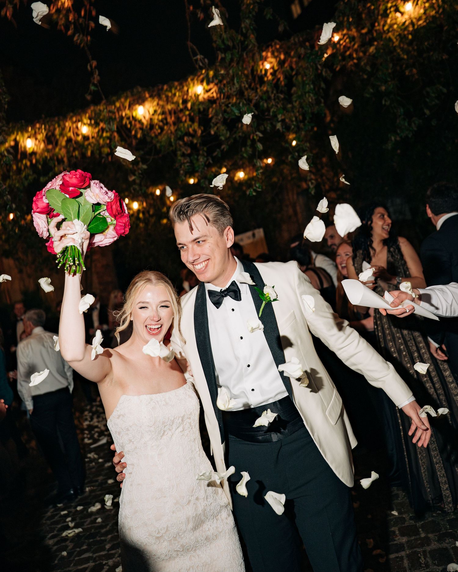 Joyful newlyweds celebrate with rose petal toss, bride holding pink bouquet in white gown, groom in ivory tuxedo jacket.