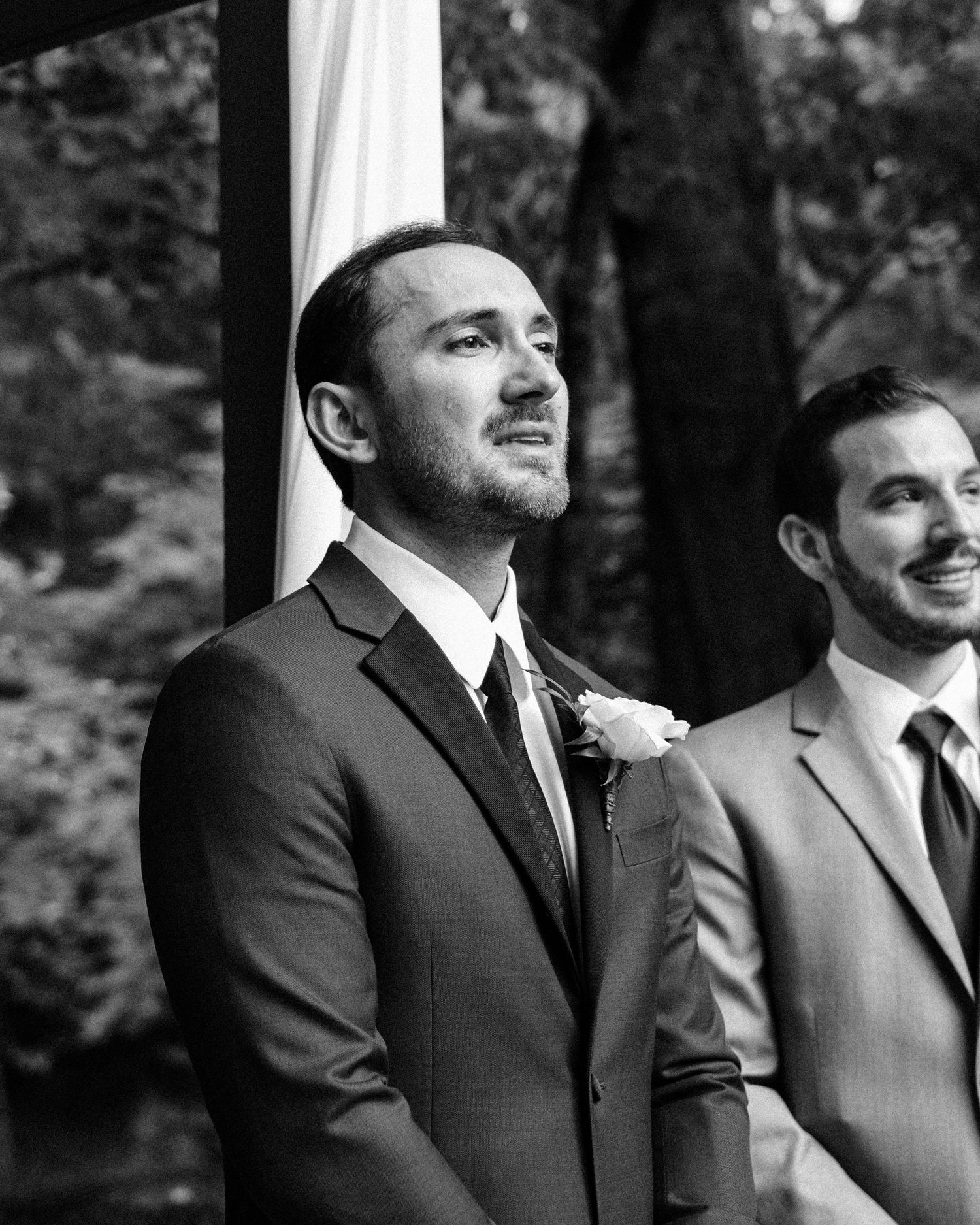 Black and white photo of a groom in a suit with boutonniere, emotional at wedding altar, best man standing beside him.