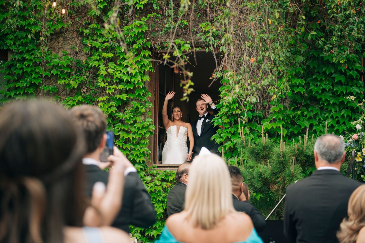 Bride and groom celebrate at outdoor wedding ceremony framed by lush green ivy-covered wall.