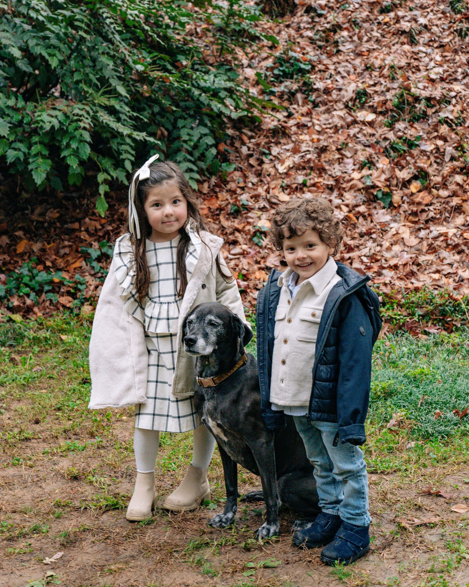 Two young children in dressy outfits pose with a black Labrador dog outdoors among autumn foliage.