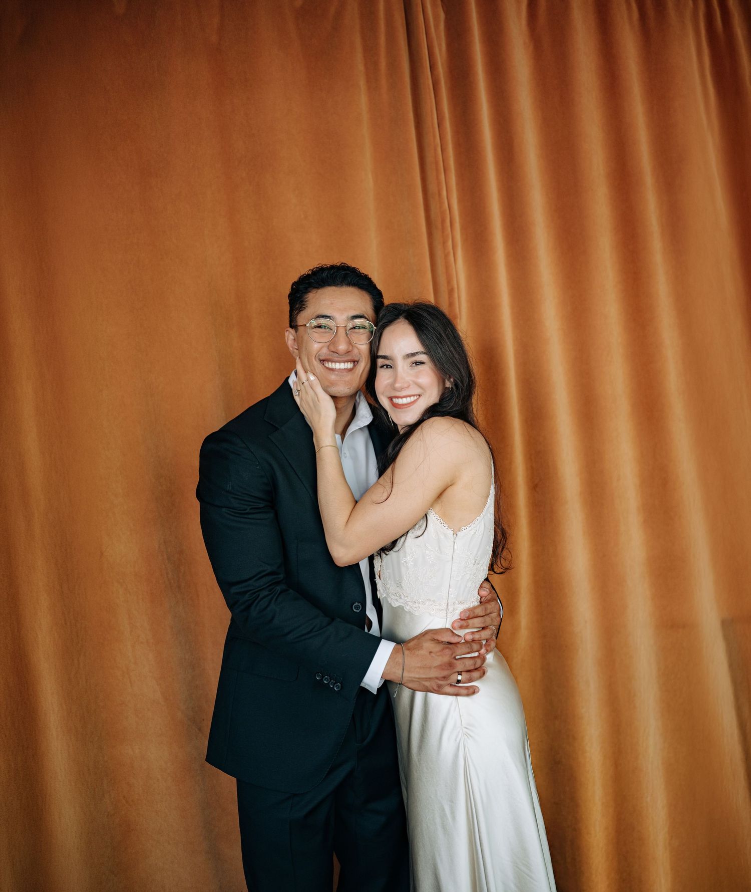 A smiling couple in formal attire embraces in front of a warm orange velvet curtain backdrop.