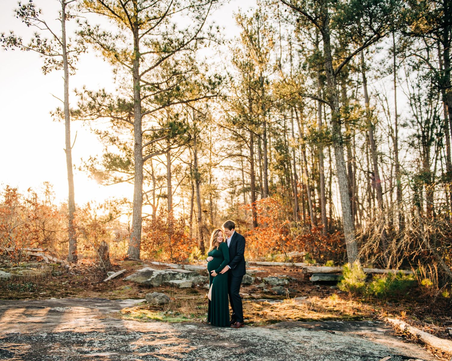 Couple embracing in a sunlit autumn forest with tall pine trees and golden foliage during an outdoor engagement session.