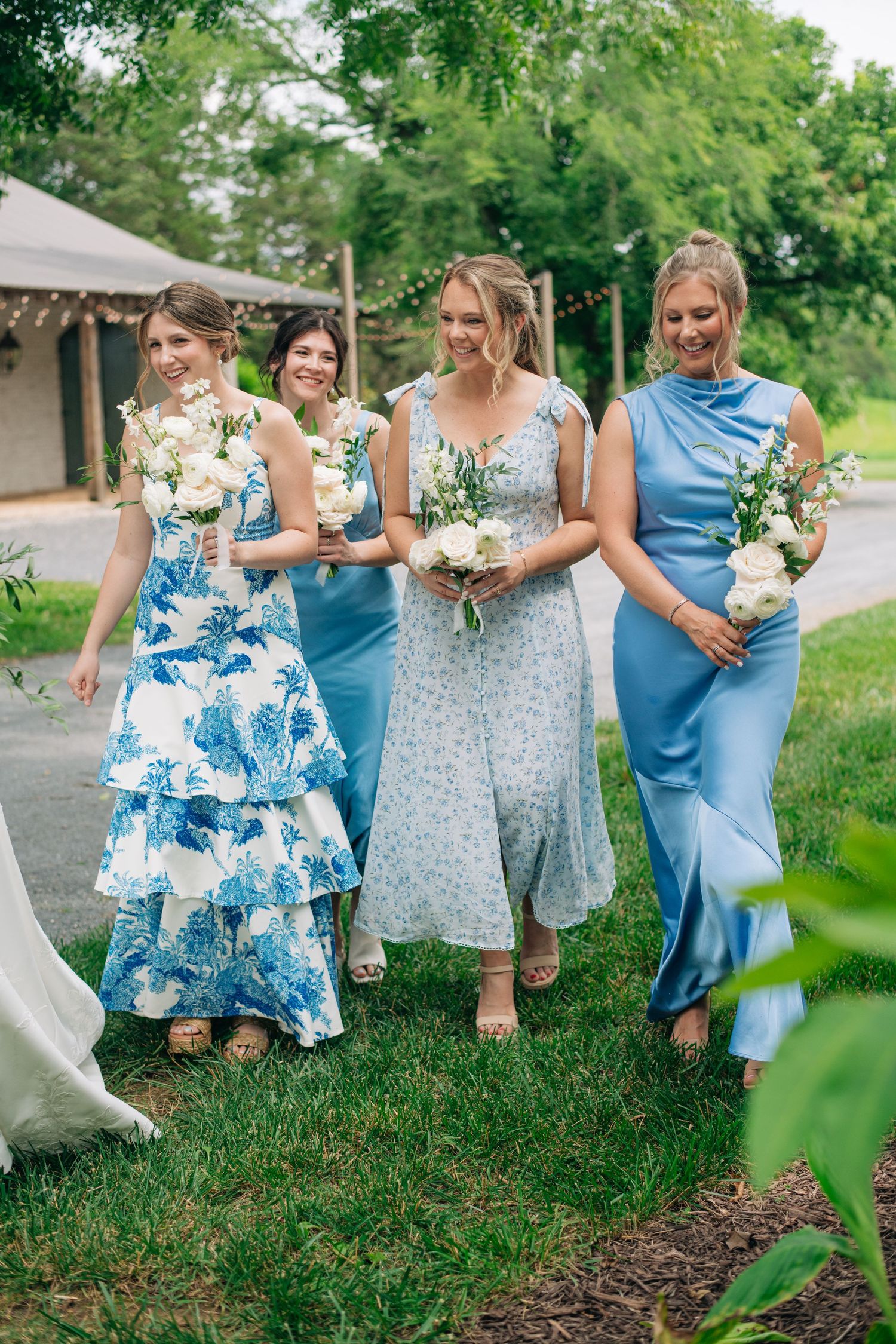 Four bridesmaids in blue dresses walk outdoors holding white bouquets at a wedding celebration.