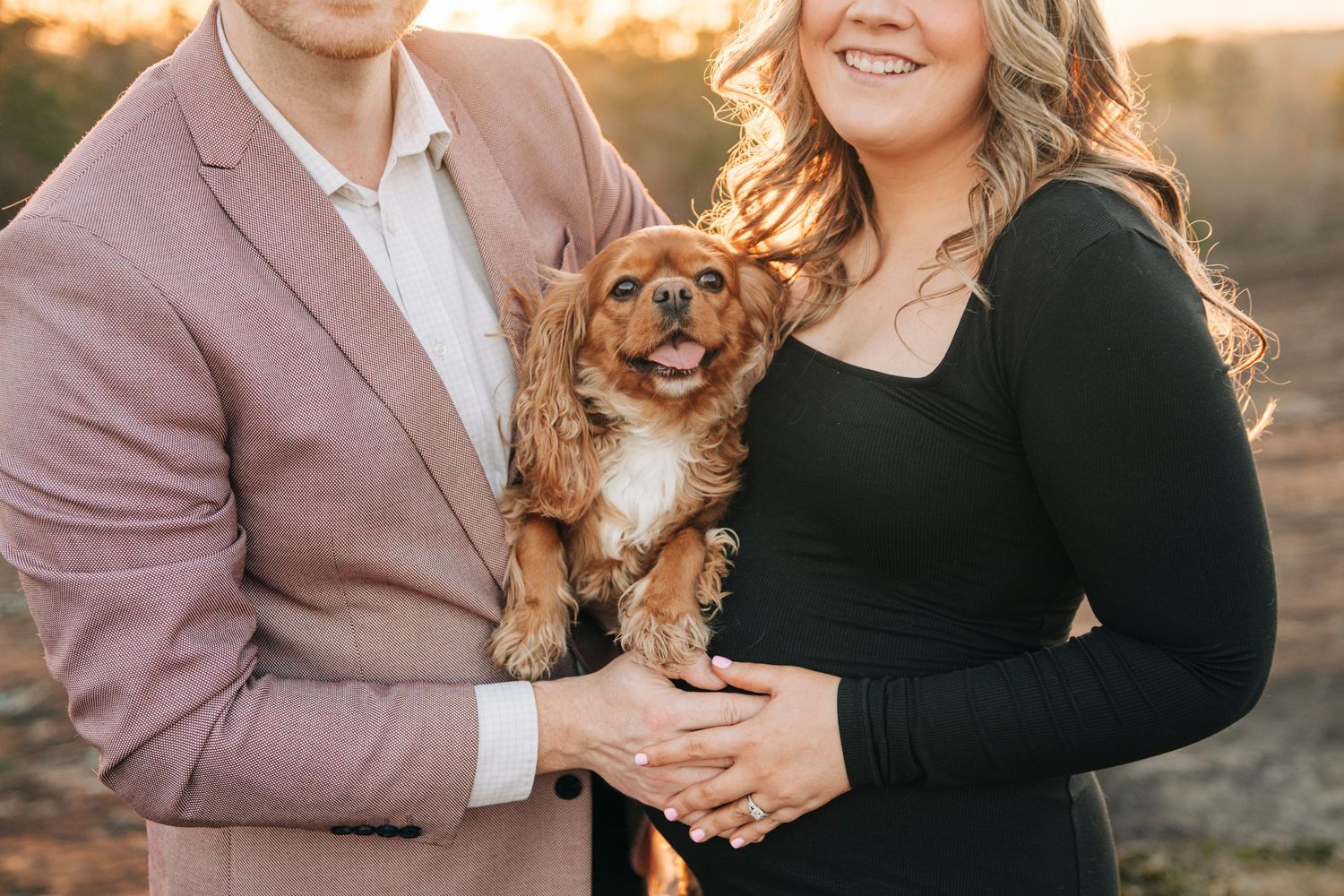 Smiling couple holding an adorable Cavalier King Charles Spaniel dog outdoors during golden hour engagement photo session.