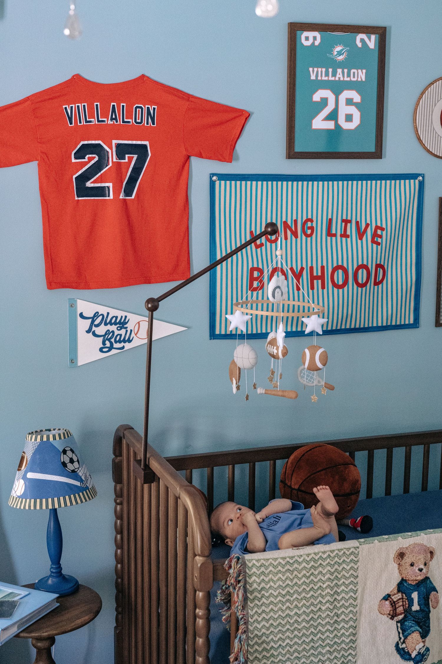 Baby nursery with sports theme, Villalon jerseys on blue wall, crib, basketball, and Long Live Boyhood banner.
