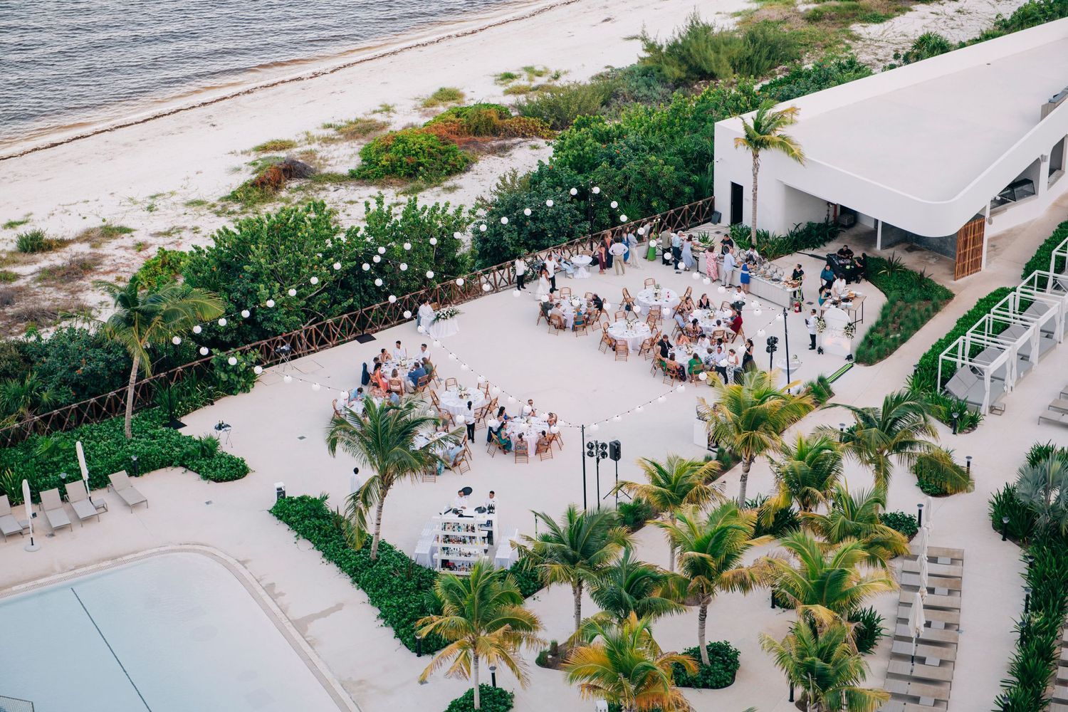 Aerial view of an elegant beachside wedding reception with round tables, palm trees, and ocean views.