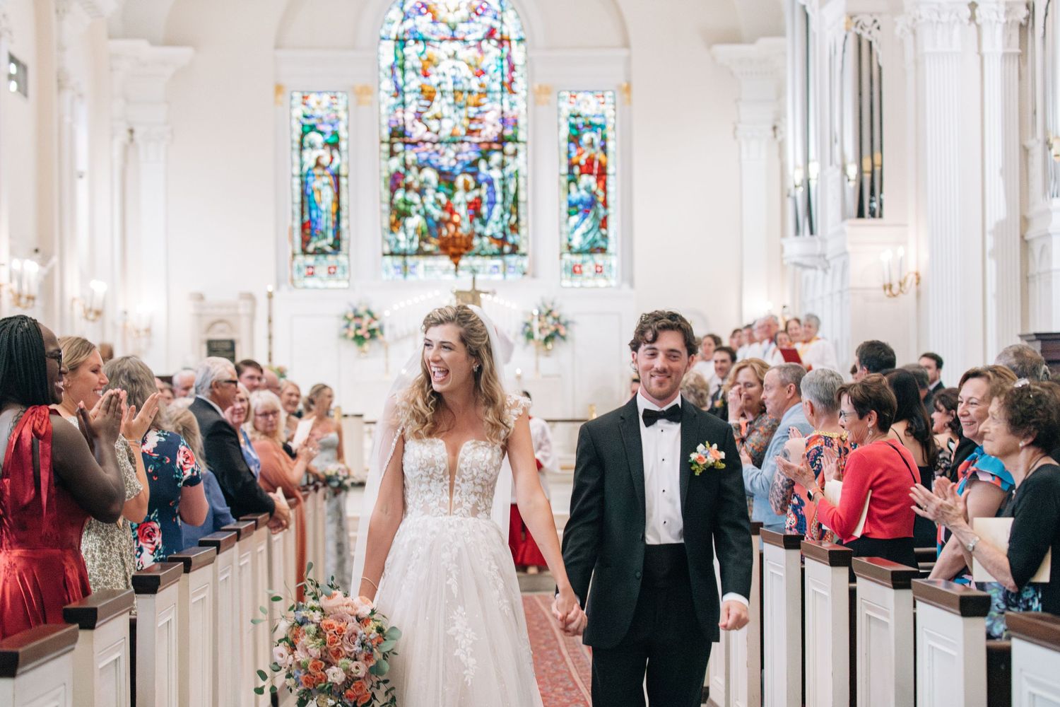 Bride and groom walk down church aisle after wedding ceremony, surrounded by cheering guests and stained glass windows.