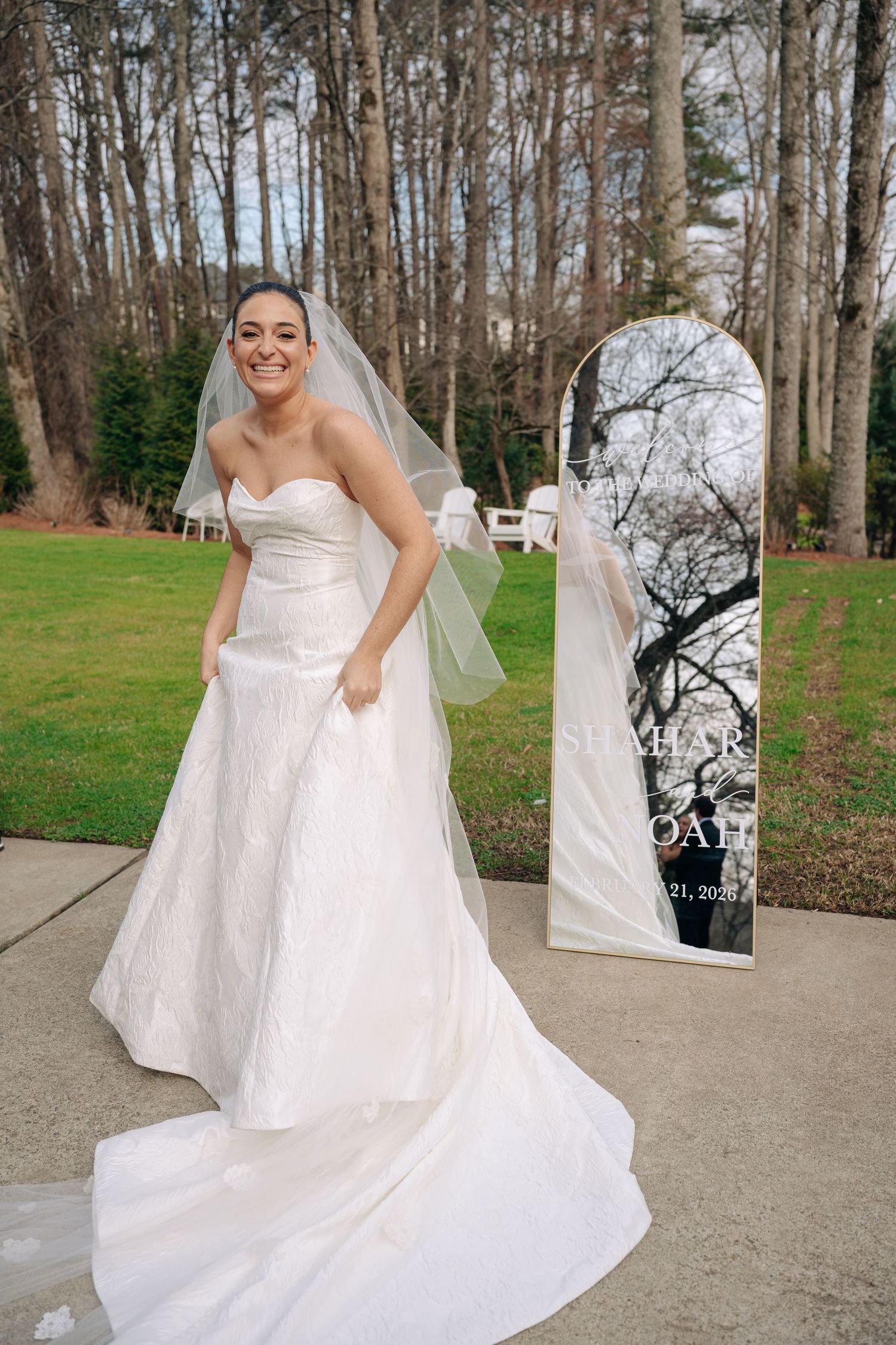 Smiling bride in strapless lace gown with veil poses outdoors near personalized wedding mirror.