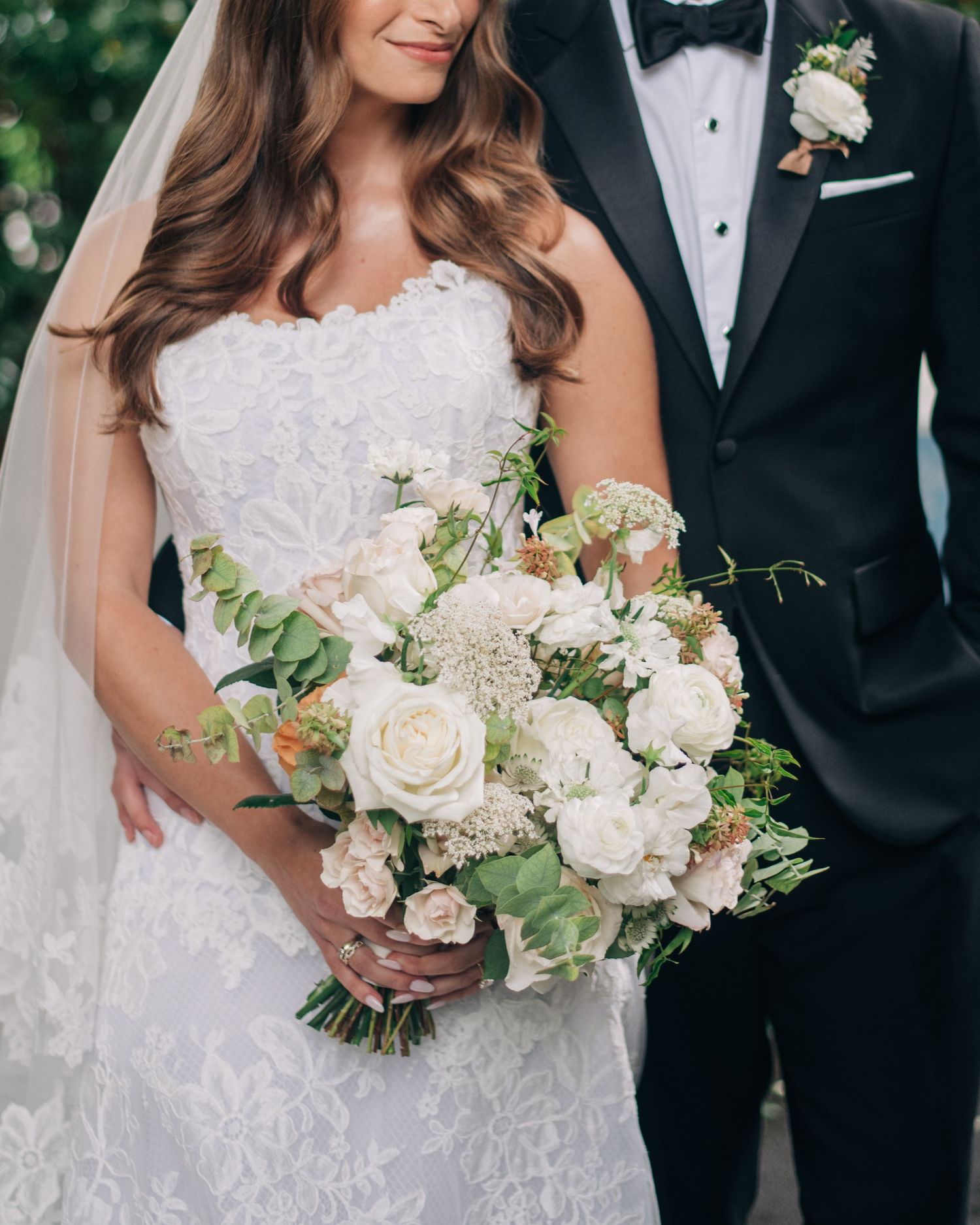 Bride in lace wedding gown holds white rose bouquet beside groom in classic black tuxedo in Highlands, NC wedding.
