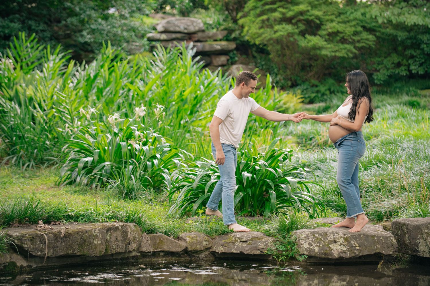 Couple standing on stepping stones by a pond in a lush green garden, woman visibly pregnant, holding hands.