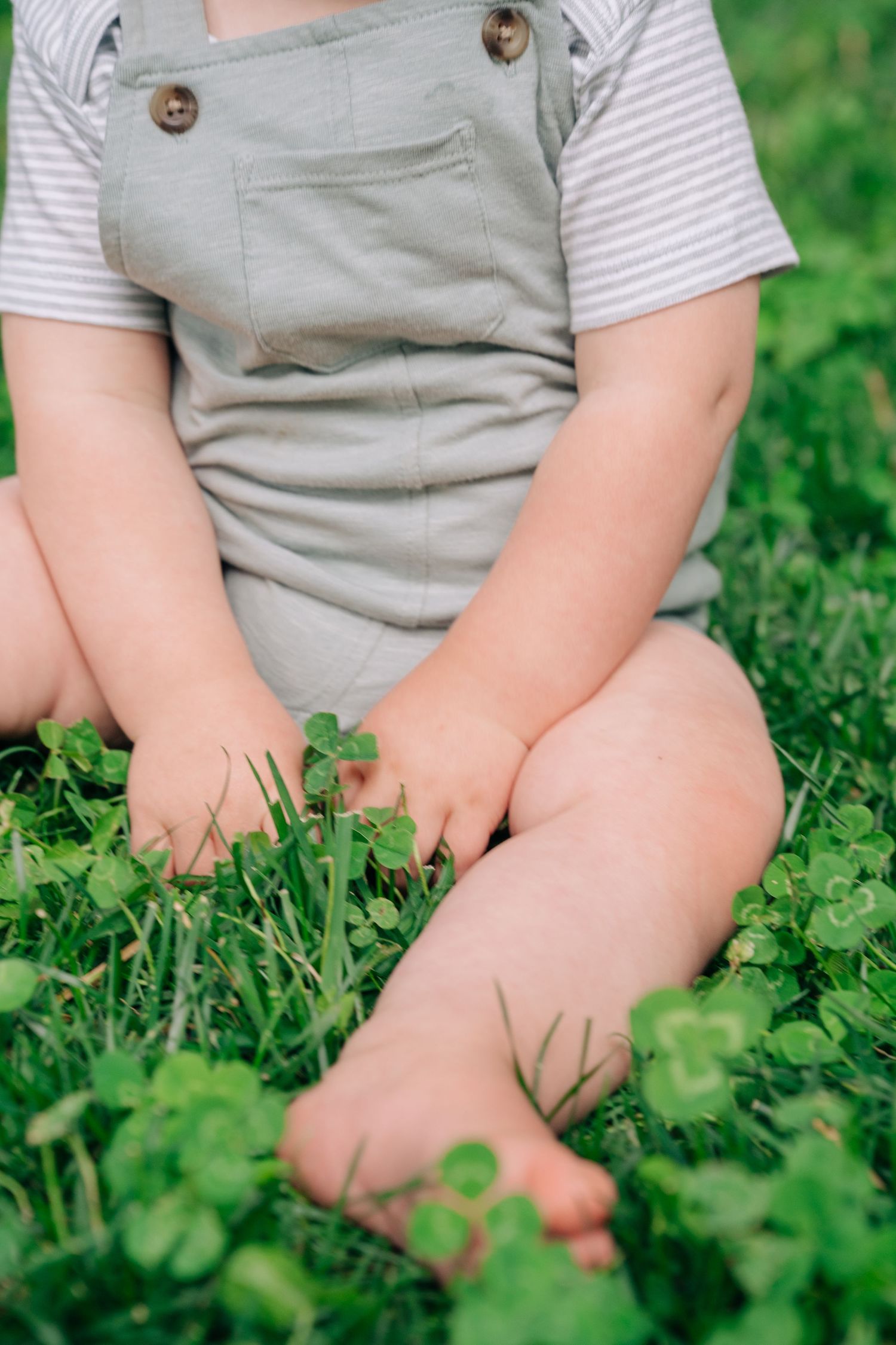Baby sitting barefoot in green clover grass wearing sage overalls and striped shirt.