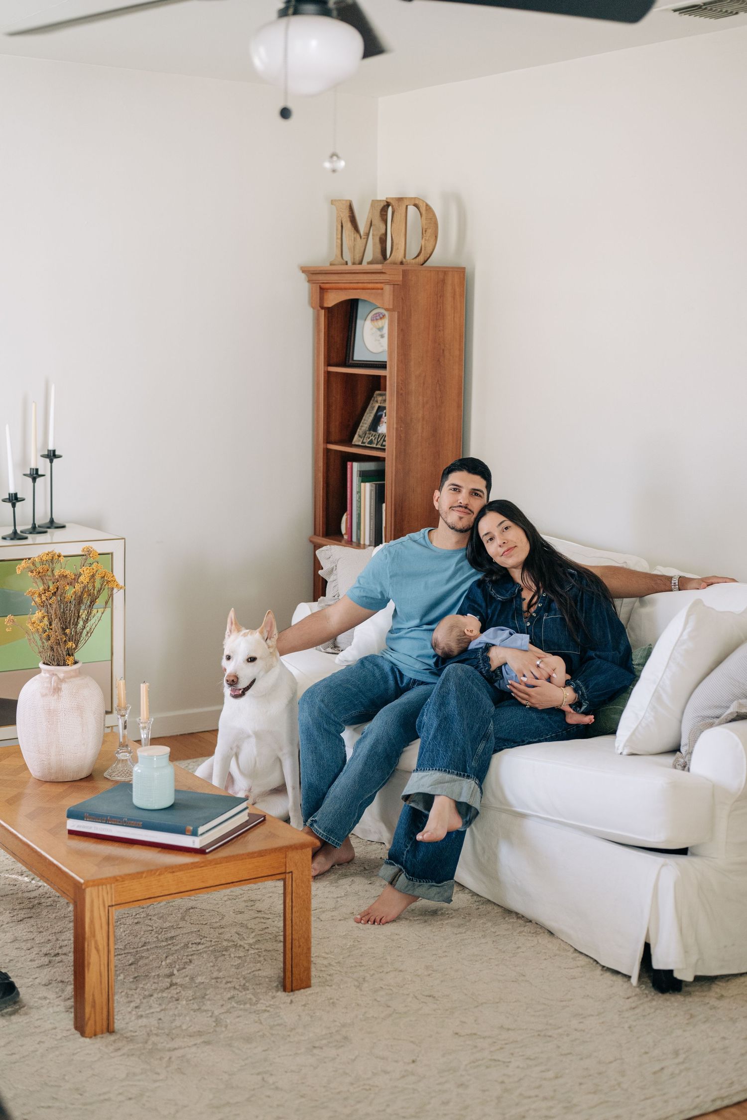 A couple relaxes on a white sofa with their newborn baby and white dog in a cozy living room.