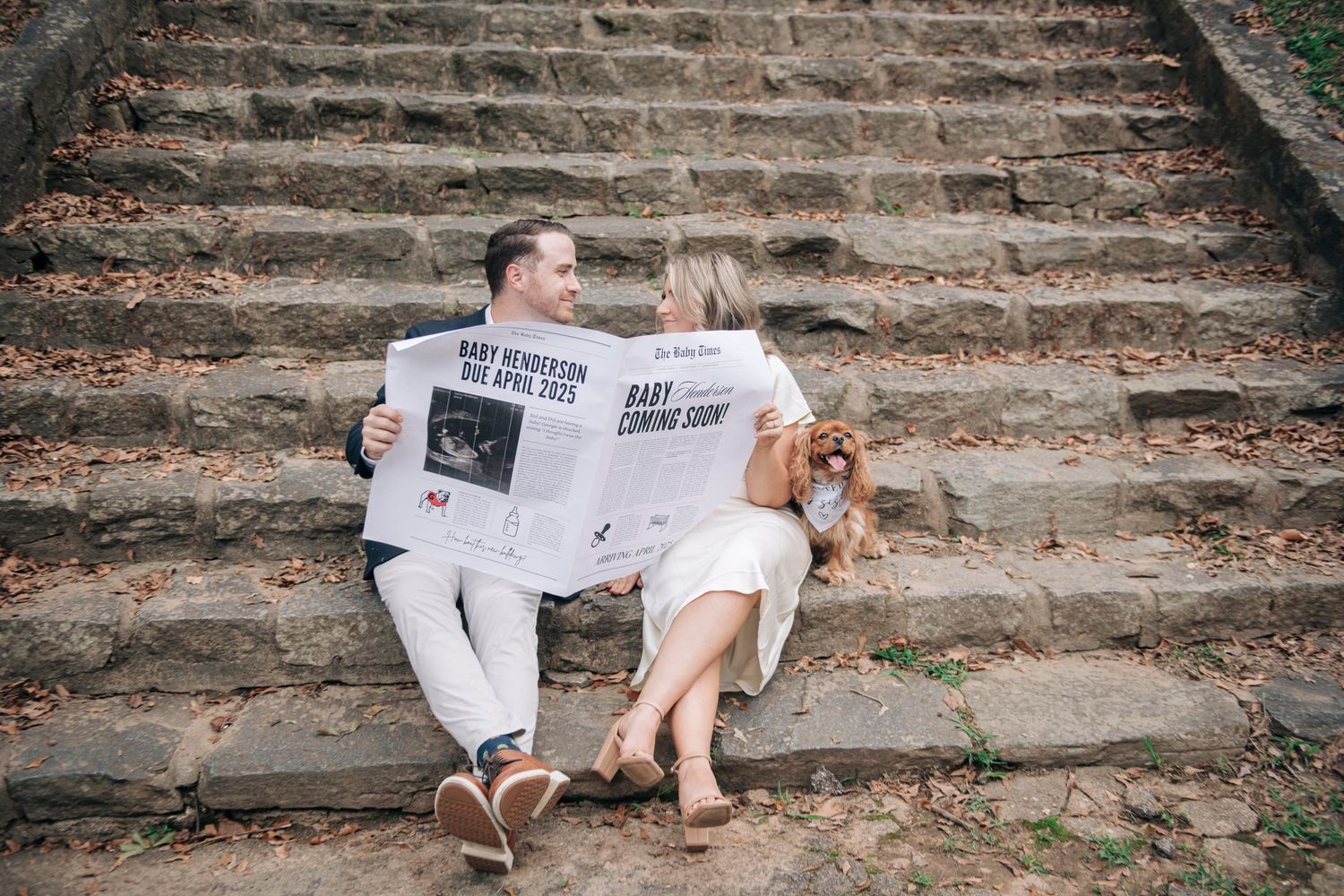 Couple sitting on stone steps holding baby announcement newspapers, pregnancy reveal photo shoot outdoors.