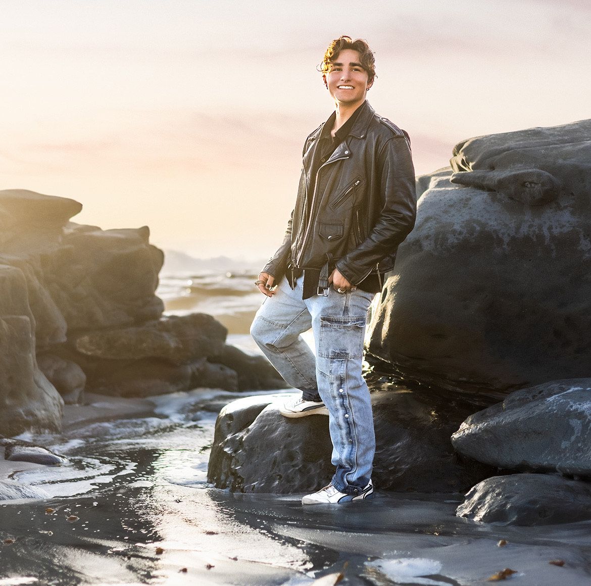 High School Senior Boy with black leather jacket and jeans posing for his senior photo at the beach in La Jolla