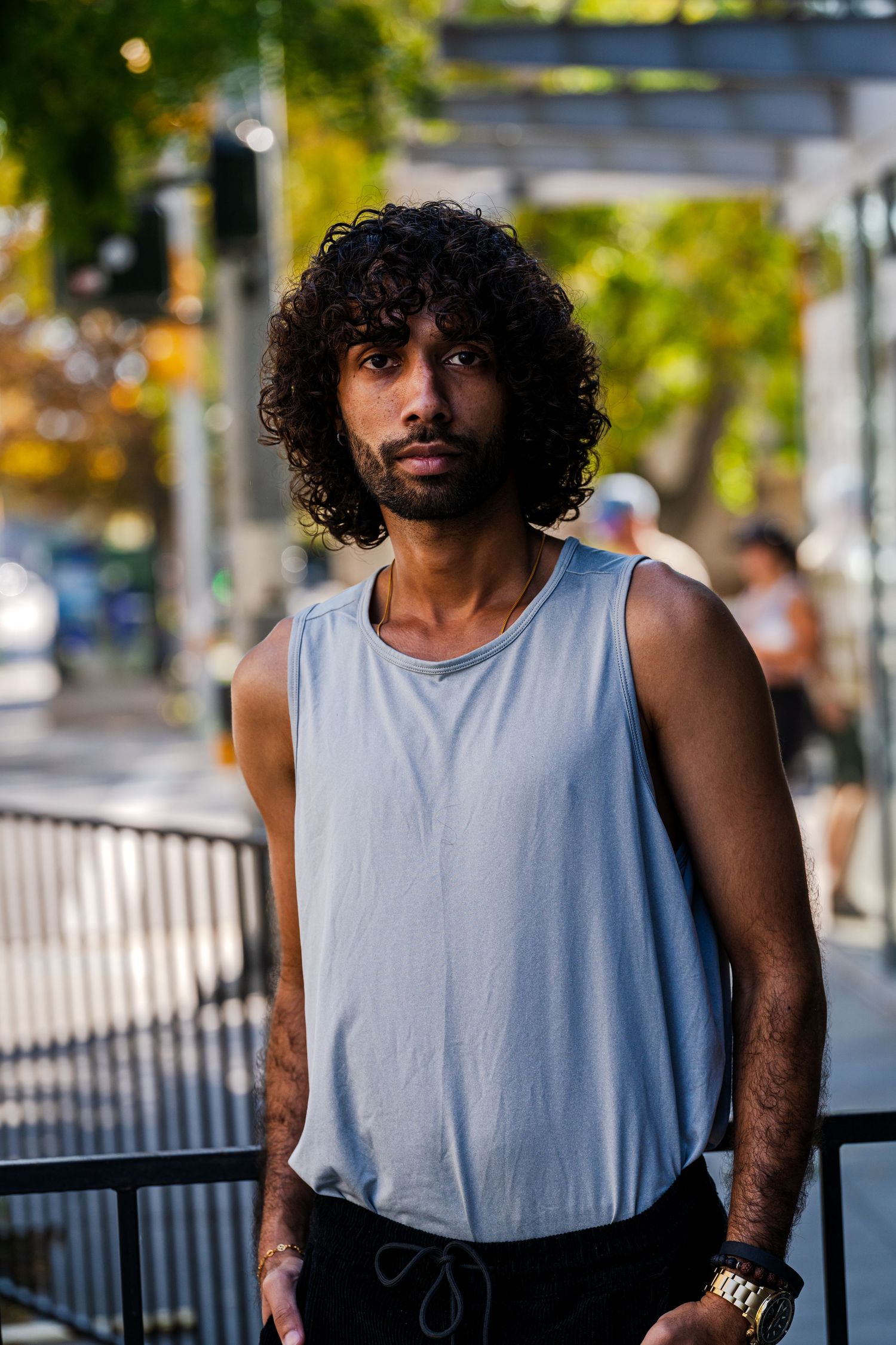 Person in gray tank top standing outdoors with curly hair and urban background.