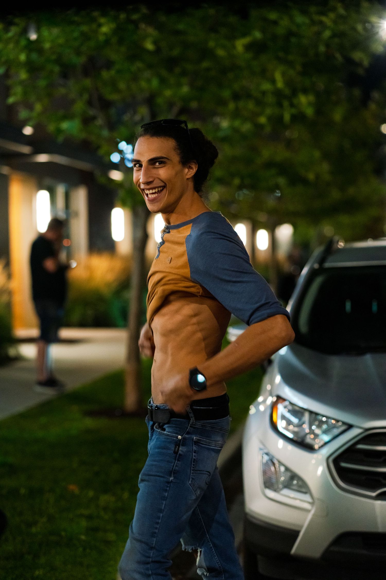 Person in brown and blue baseball shirt smiling at night with car and trees in background.