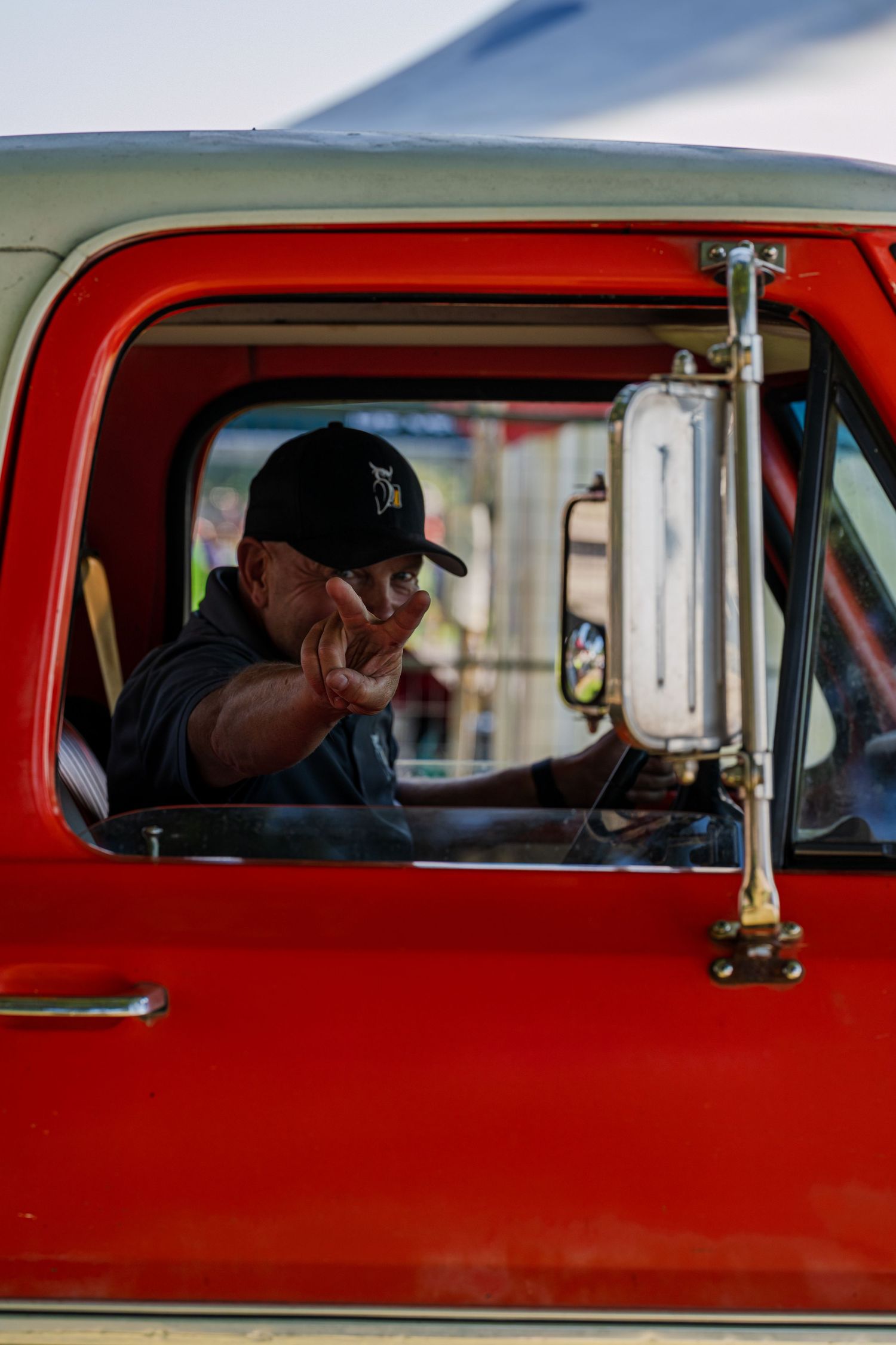 Person in baseball cap gesturing from window of vintage red truck.