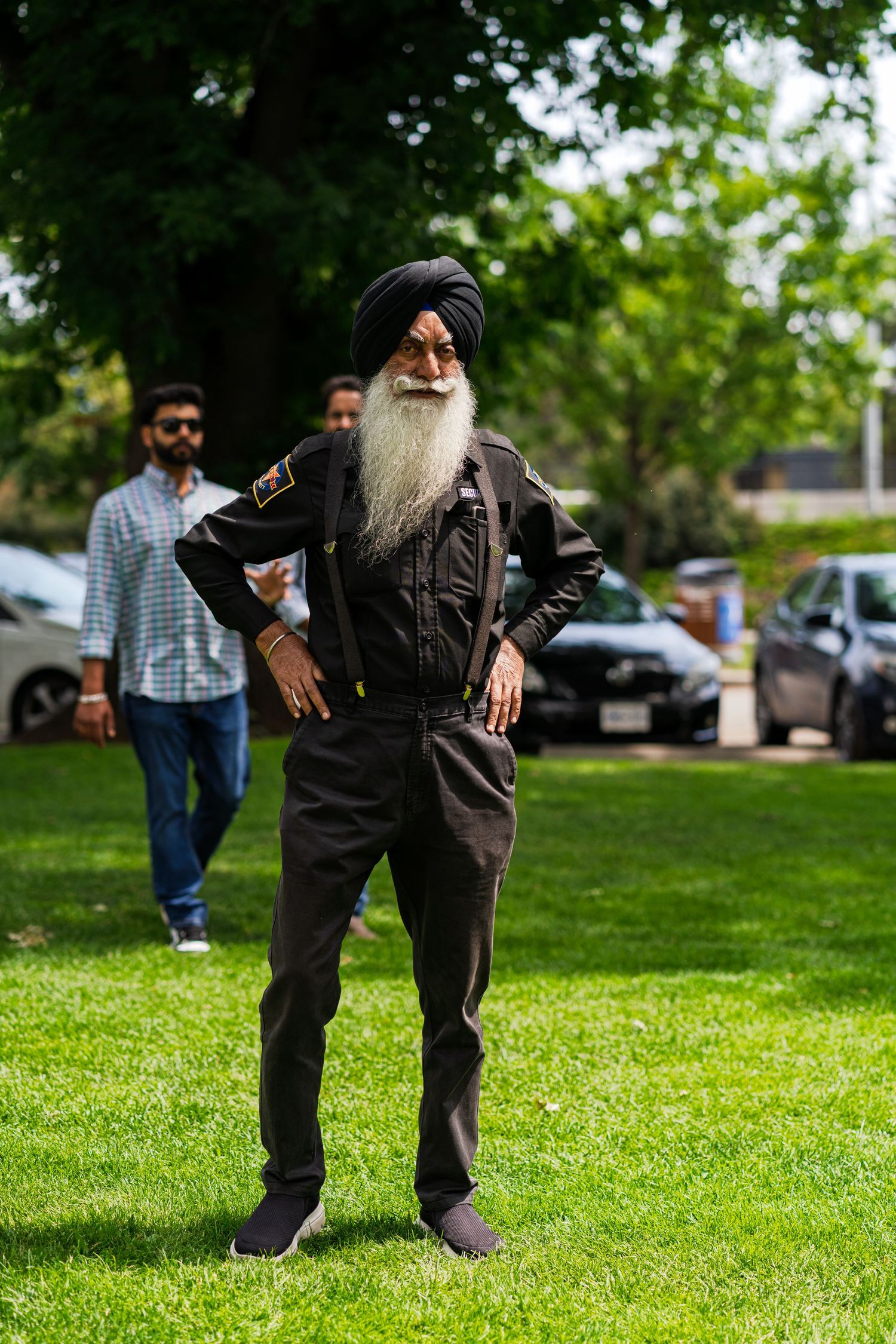 Person in black outfit and turban standing on grass with parked cars in background.