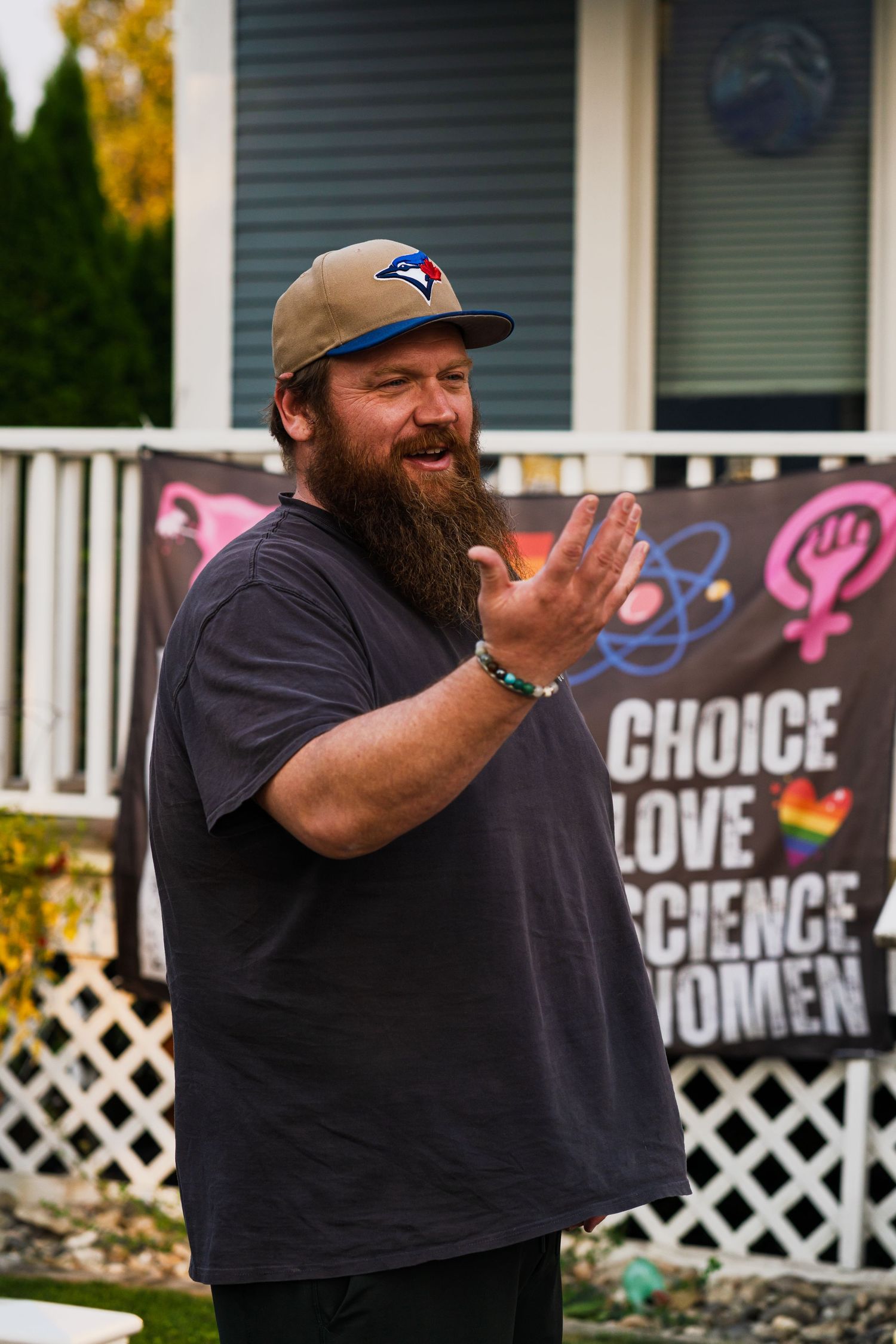Man in black t-shirt and Toronto Blue Jays cap gestures expressively while standing outside a house with lattice fencing.