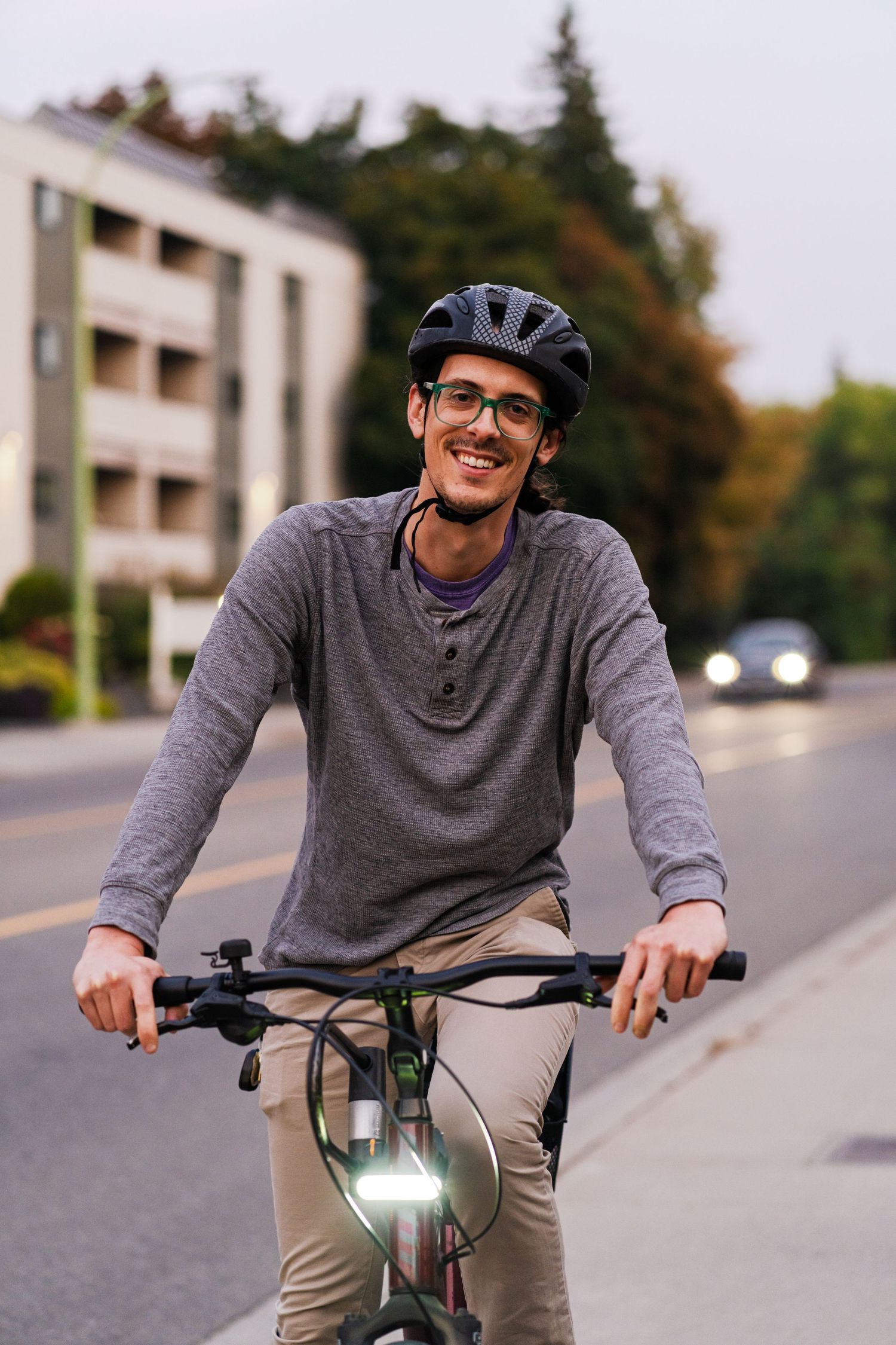 A cyclist in a grey sweater and baseball cap riding an electric bike on a suburban street.