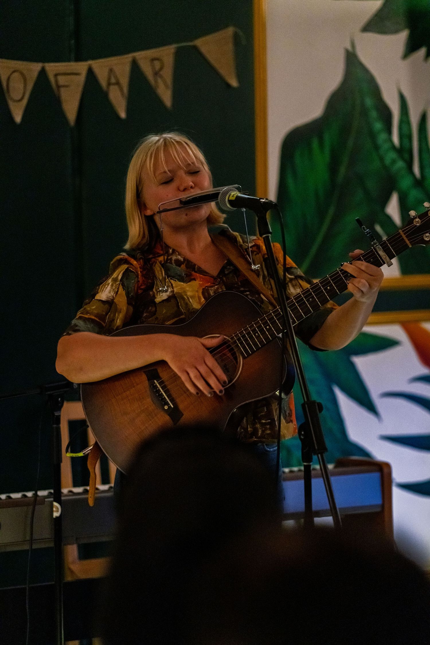 Musician performing on acoustic guitar in a dimly lit venue with decorative foliage in the background.