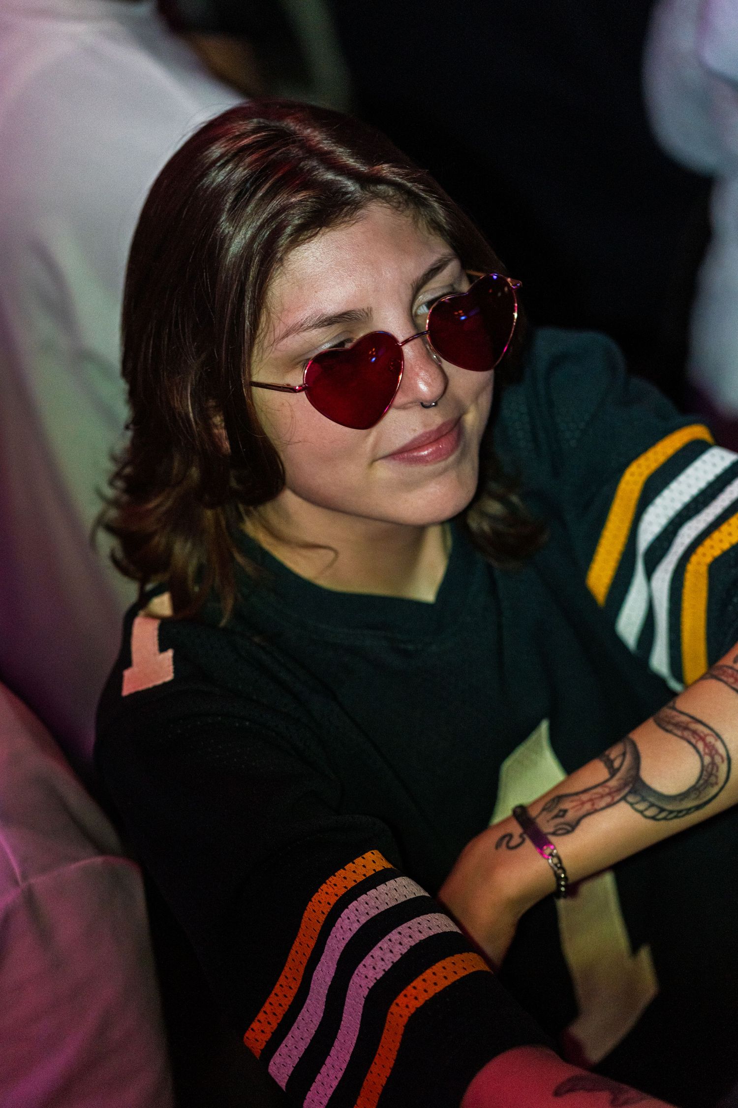 Person wearing heart-shaped sunglasses and black shirt with visible arm tattoo in dark indoor setting.