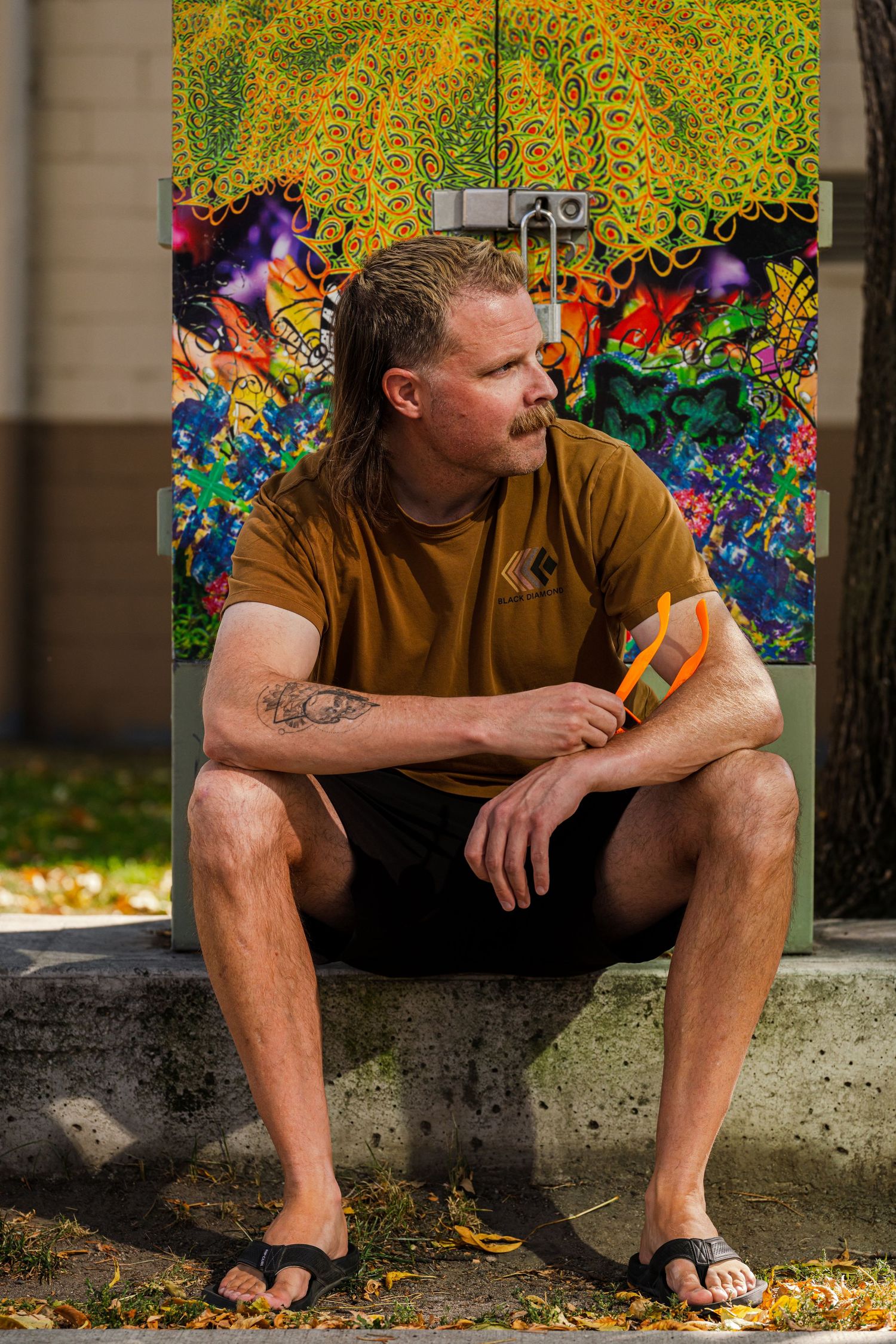 Person sitting on concrete ledge against colorful psychedelic mural wearing brown t-shirt and shorts.